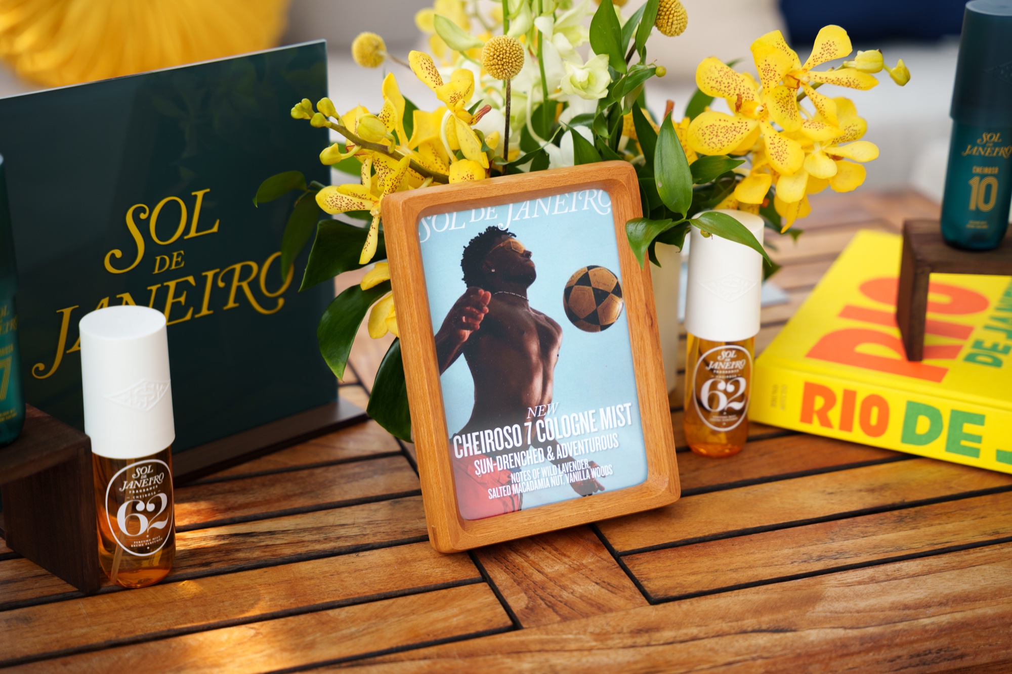 Sol de Janeiro fragrance bottles and a framed magazine cover among yellow orchids on a wooden table