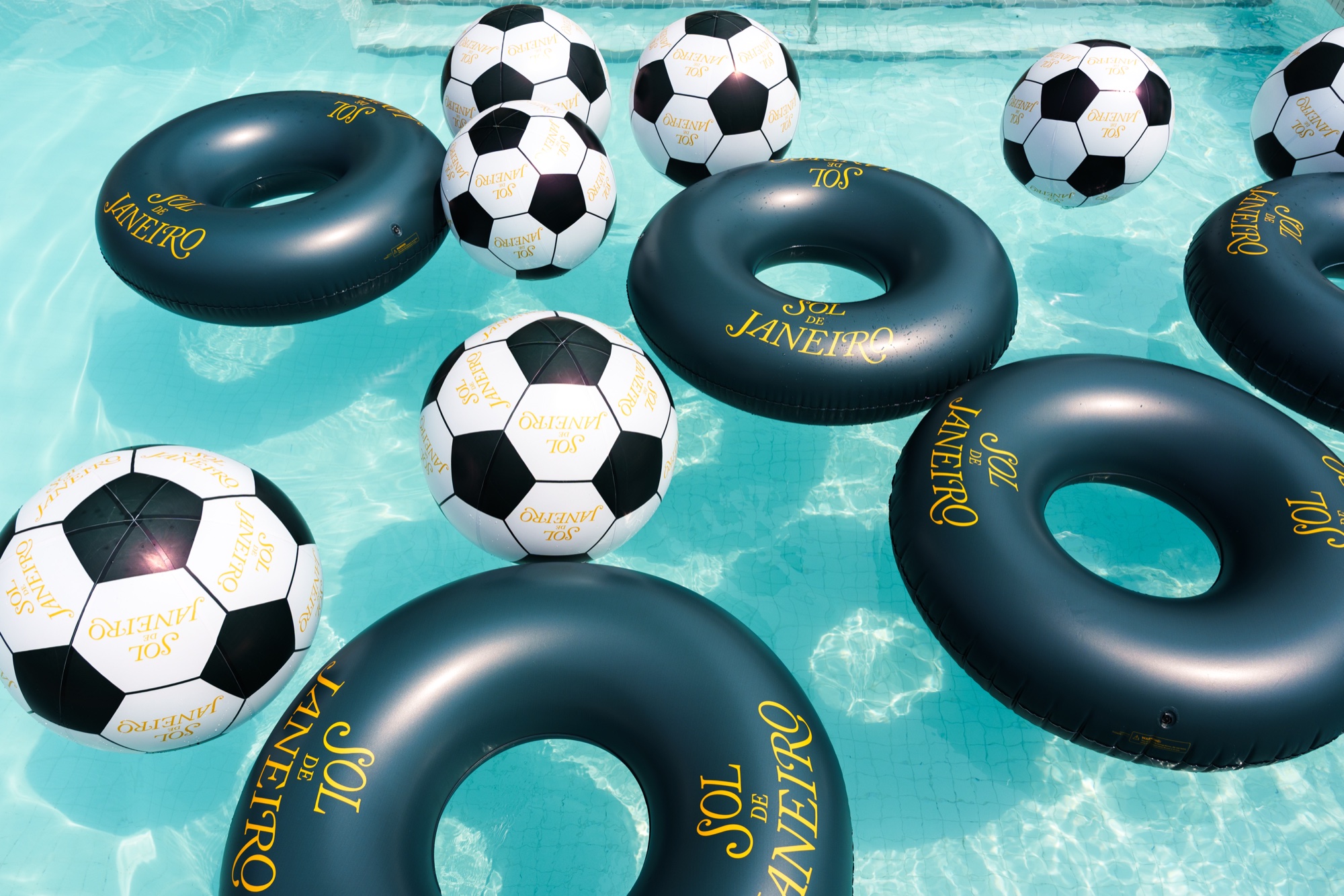 Black inflatable rings and soccer balls printed with Sol de Janeiro float on turquoise pool water