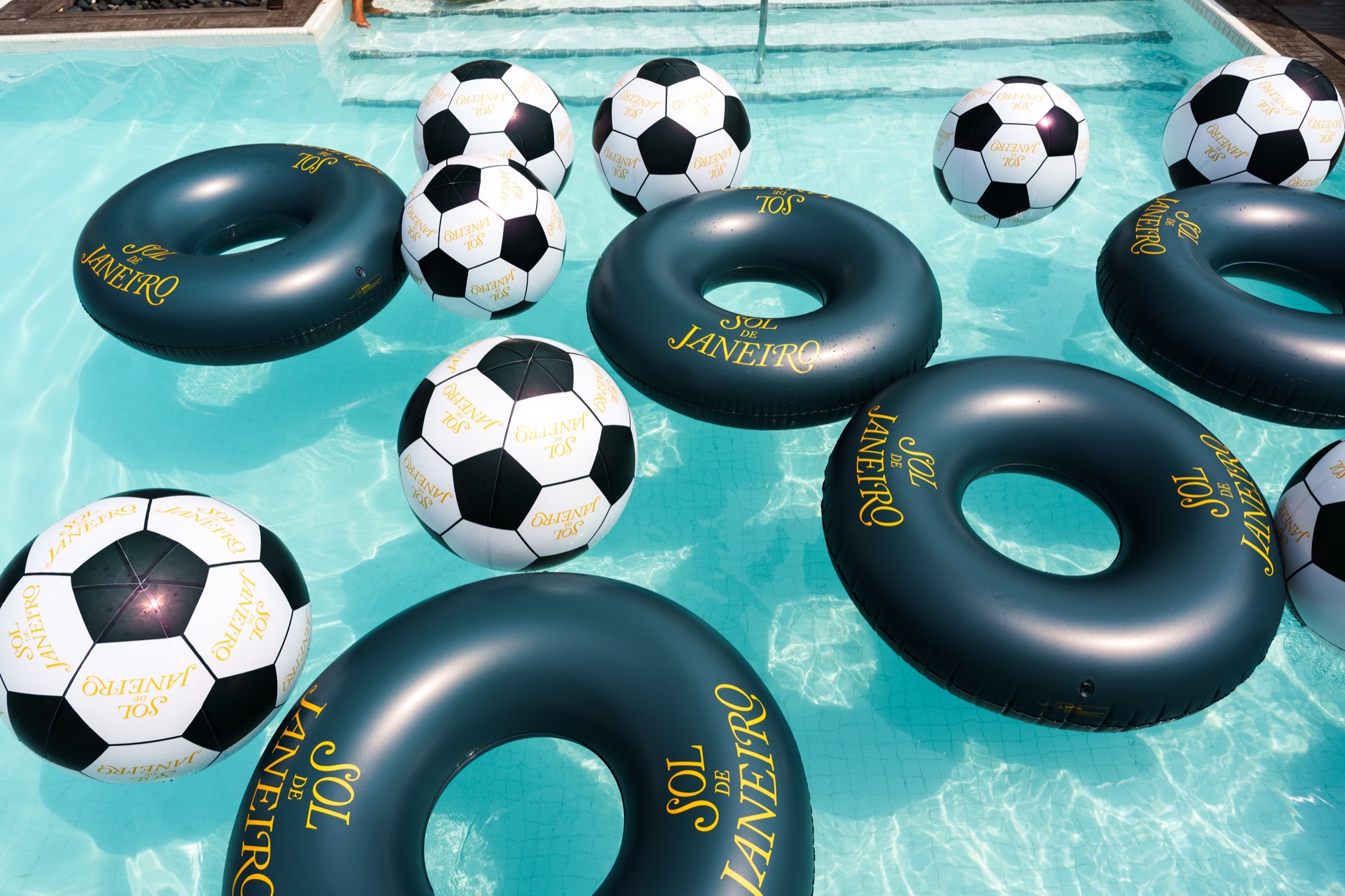 Cluster of soccer ball beach balls and green Sol de Janeiro rings floating on bright turquoise pool water