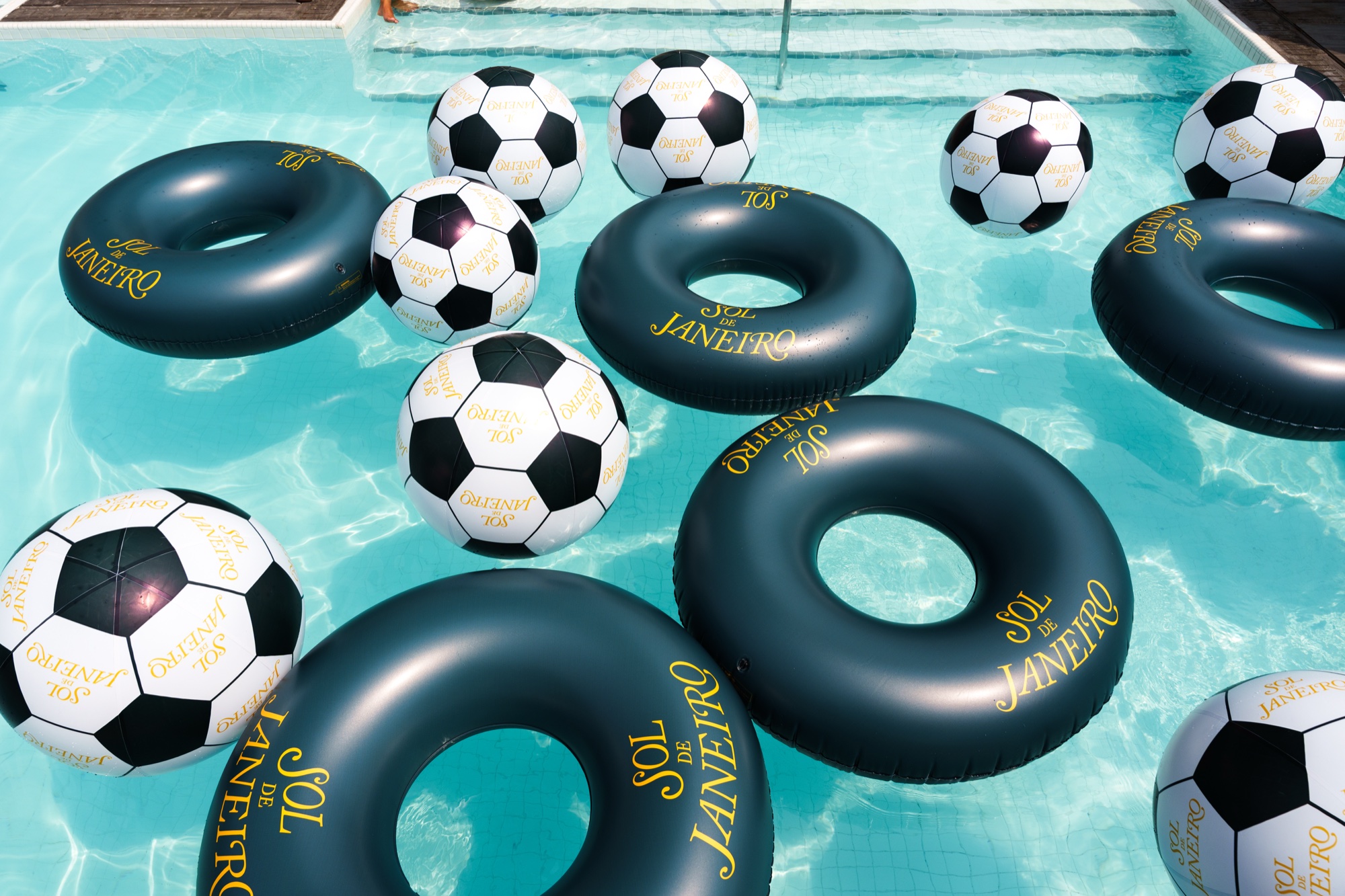 Black inflatable rings and black and white soccer balls printed with Sol de Janeiro crowd a turquoise pool