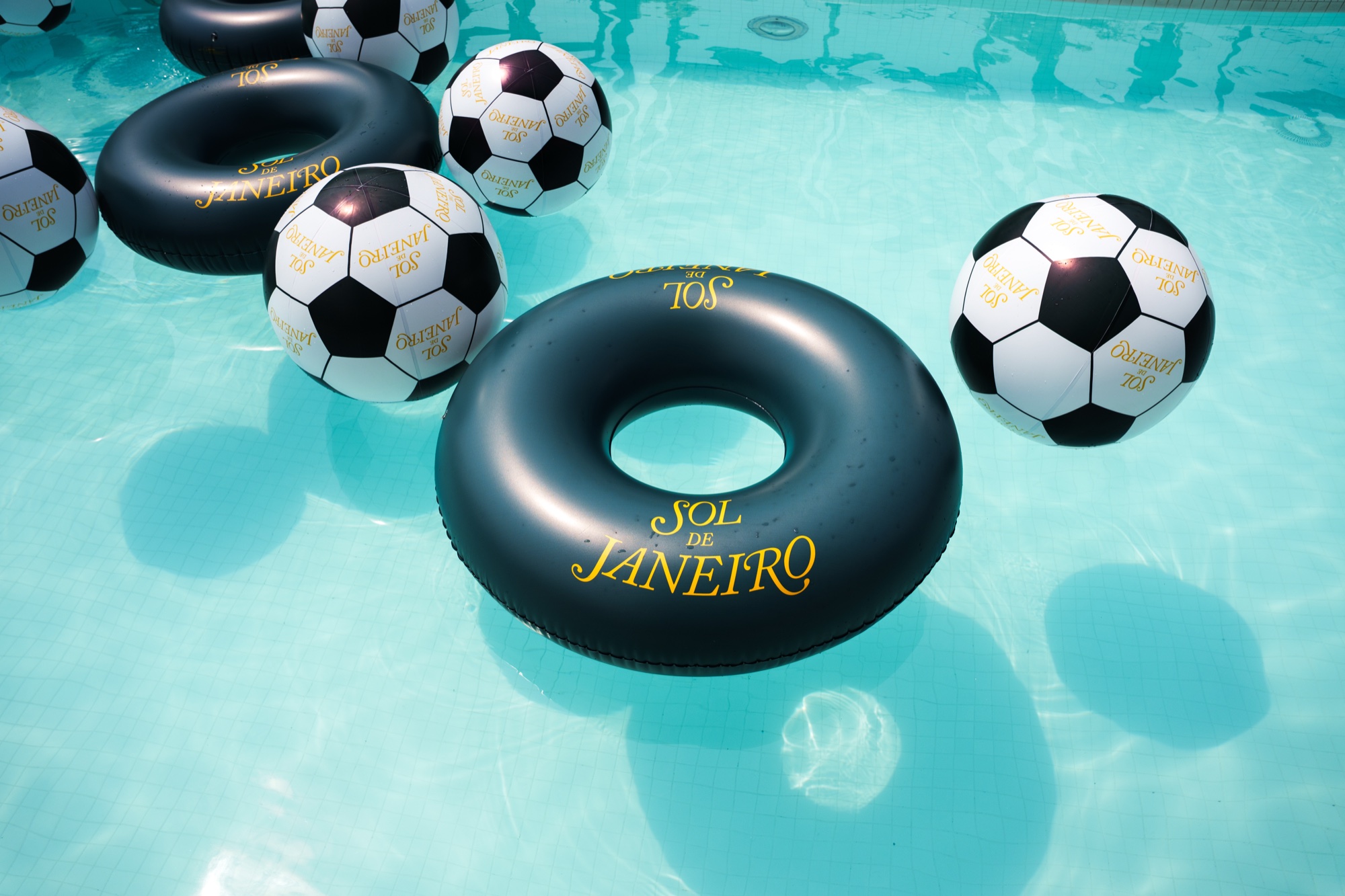 Black Sol de Janeiro pool ring floating among soccer ball beach balls in clear turquoise water