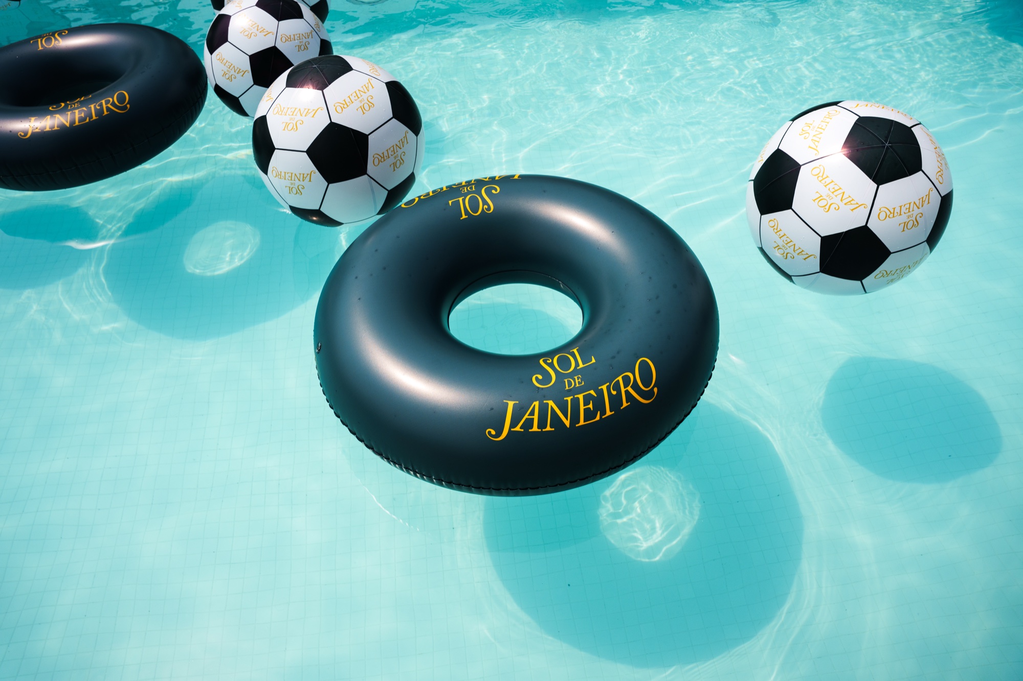 A black Sol de Janeiro pool ring floats among inflatable soccer balls on bright turquoise water
