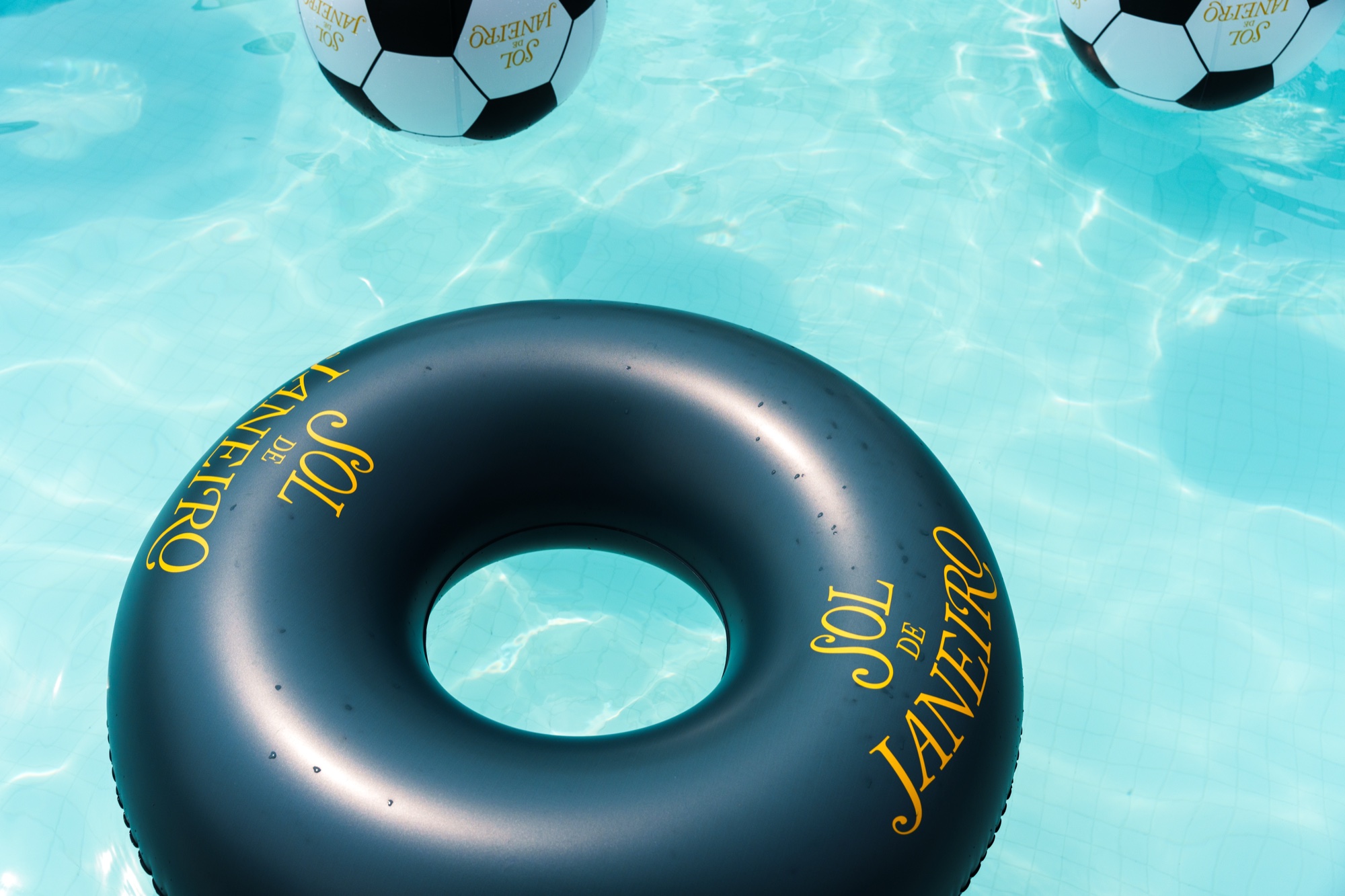 Single dark green Sol de Janeiro ring floating in clear turquoise water with two soccer balls behind