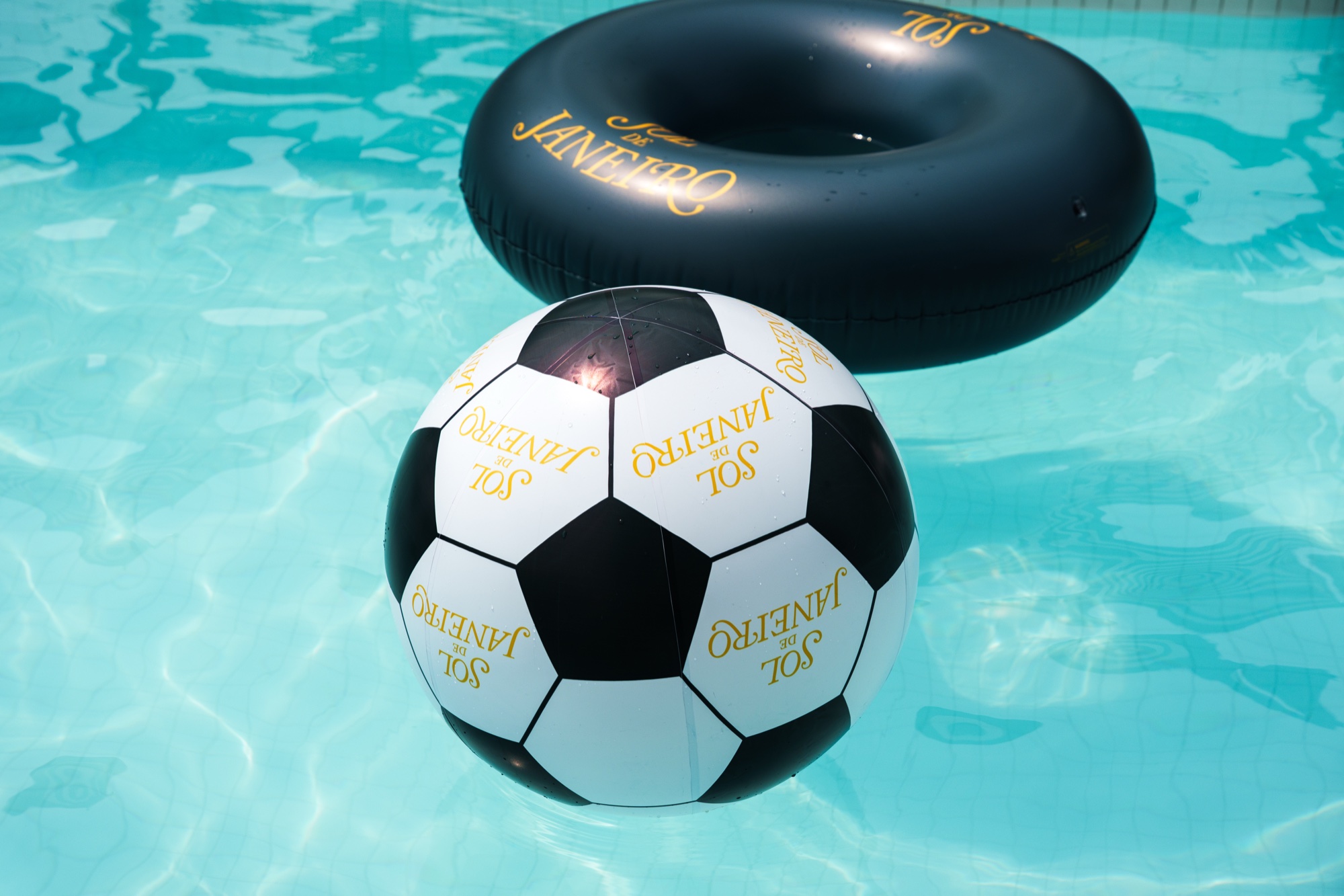 A soccer ball printed with Sol de Janeiro floats beside a black inflatable ring on rippling blue water