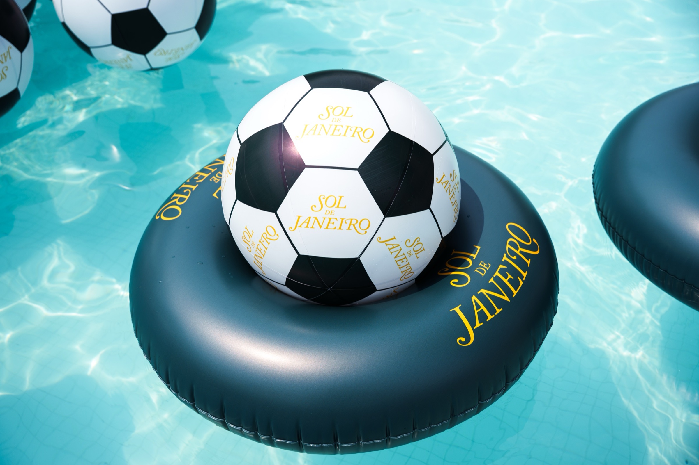 A Sol de Janeiro soccer ball beach ball resting on a navy JANEIRO inner tube in turquoise pool water