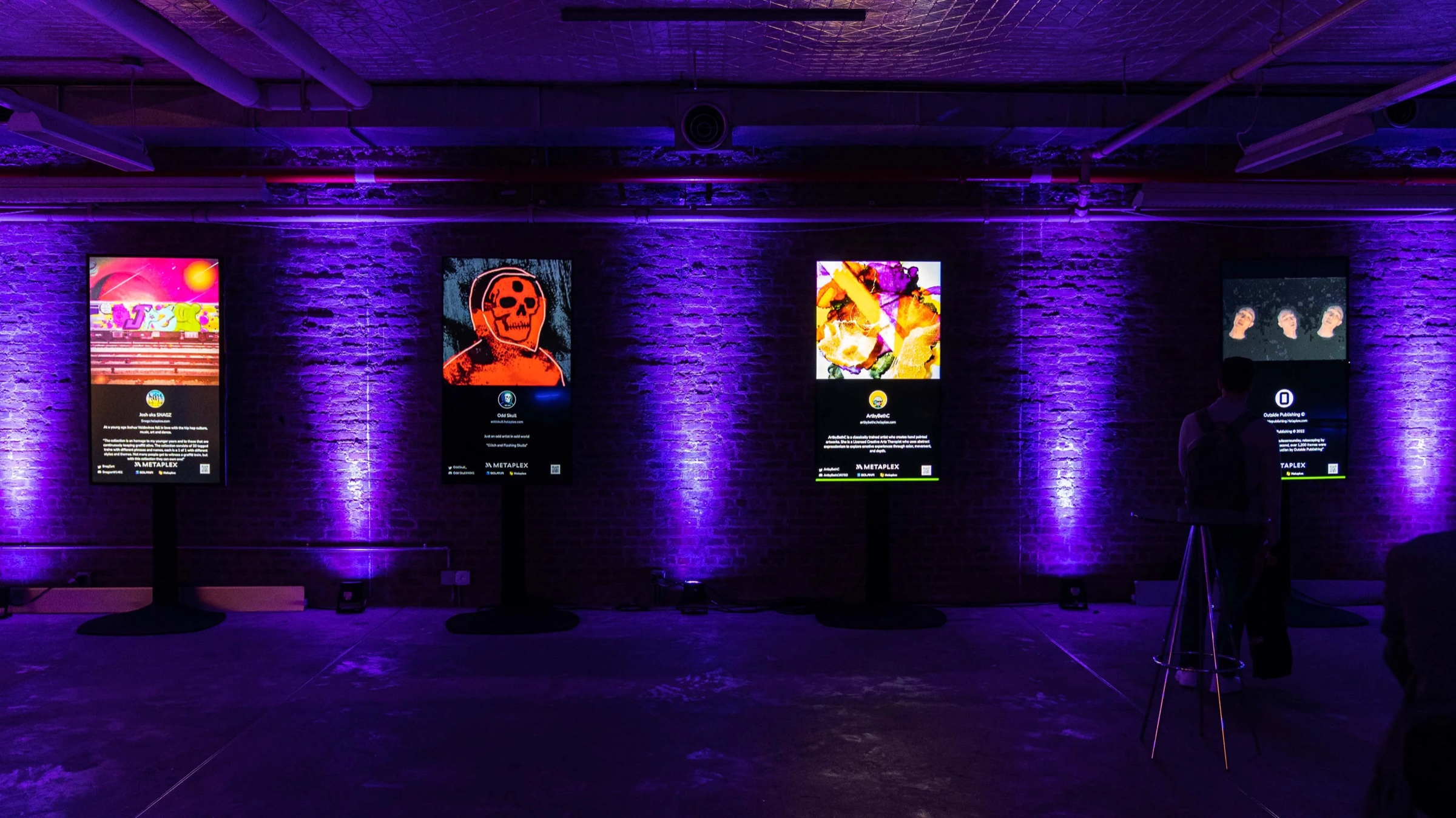 Four illuminated NFT artwork panels stand against a purple lit brick wall