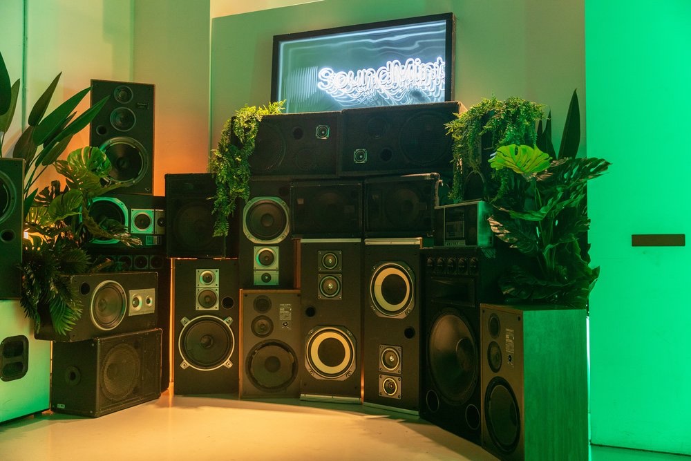 Wall of vintage speakers draped in tropical plants under a glowing SoundMint screen in green and orange light