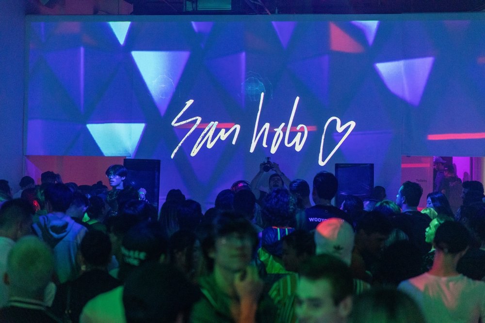 Audience faces a San Holo signature projected over purple triangle visuals at the SoundMint show