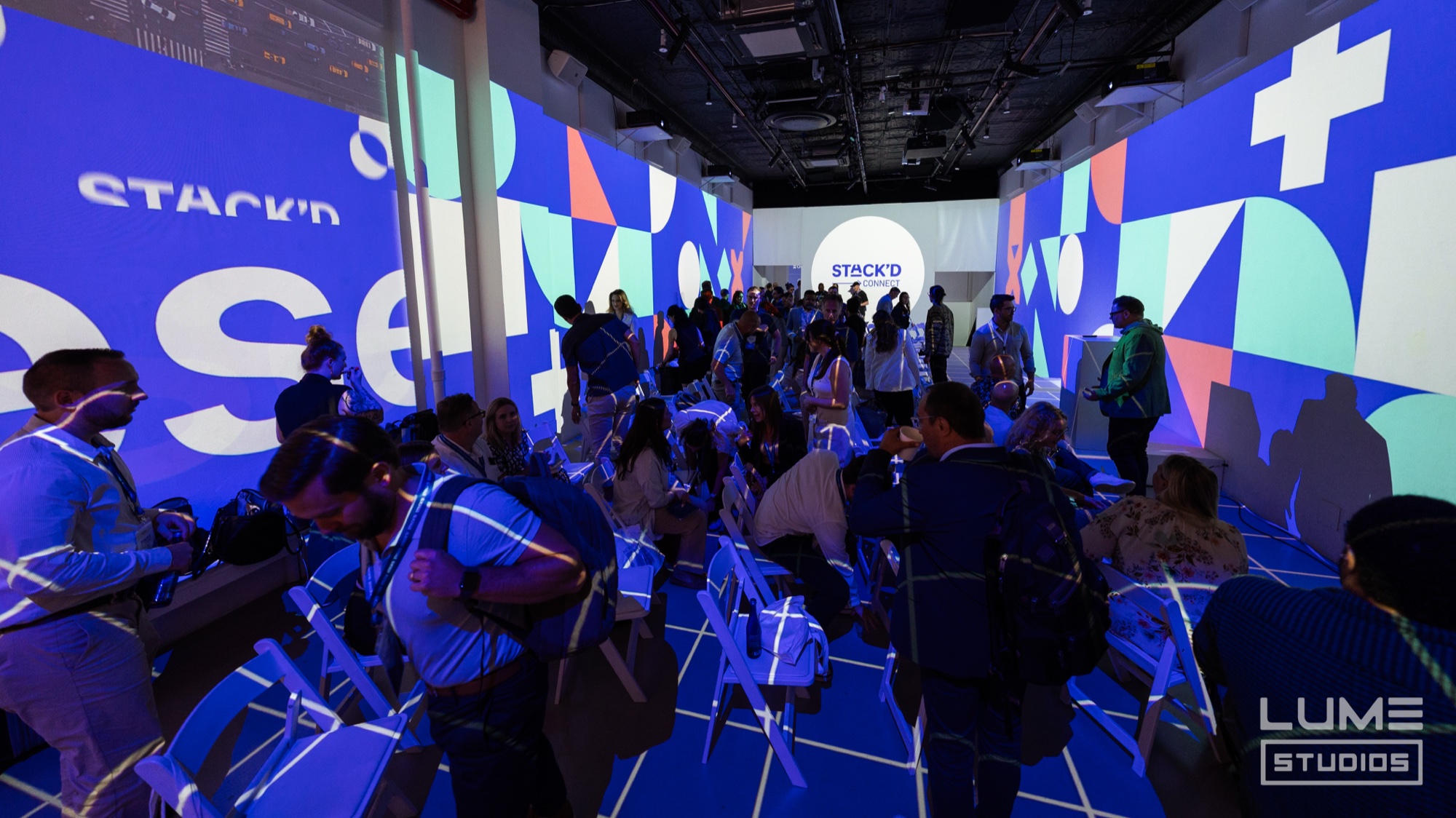 Attendees seated on white folding chairs inside Lume Studios as blue geometric Stack’d graphics wrap every wall