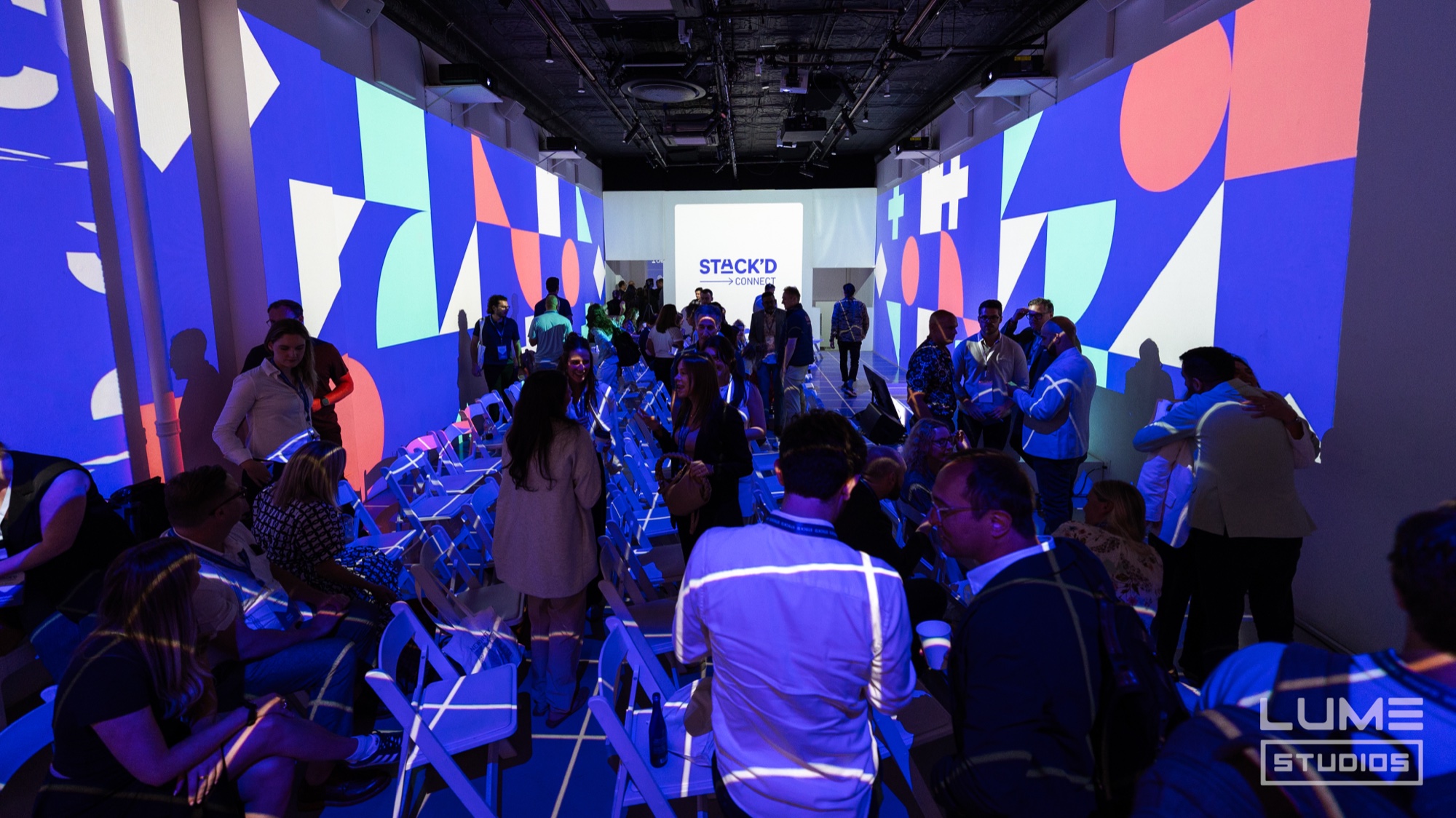 Rows of folding chairs face a Stack’d screen between walls of blue, orange and mint shapes