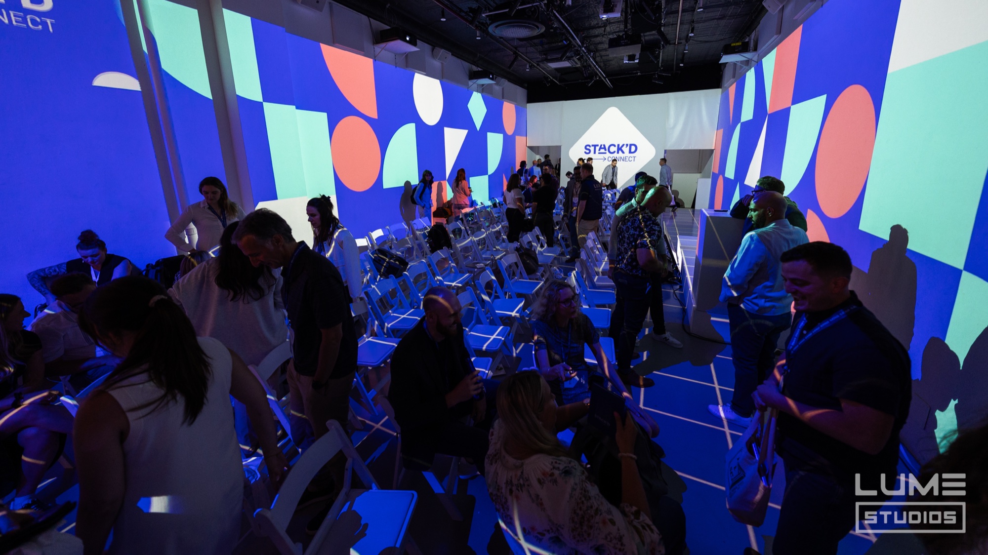 Guests find seats in a blue lit room with colourful geometric projections and a Stack’d sign ahead