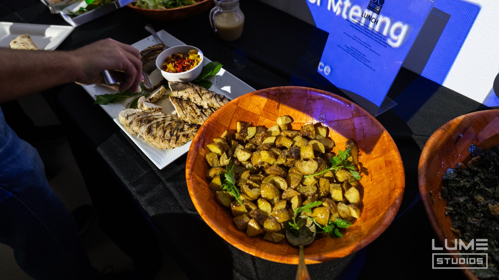 Wooden bowl of roasted potatoes with herbs beside grilled meat and salad on a black catering table
