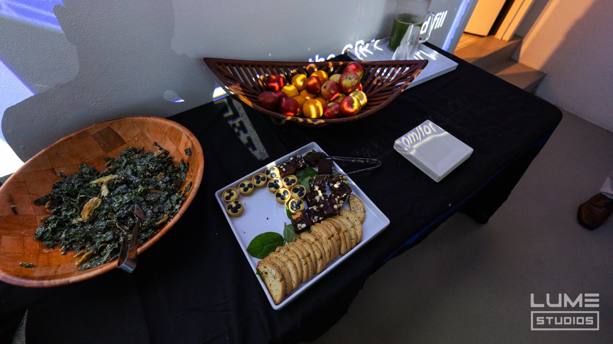 Buffet table with kale salad, cheese and cracker platter and a basket of fruit under blue projected light