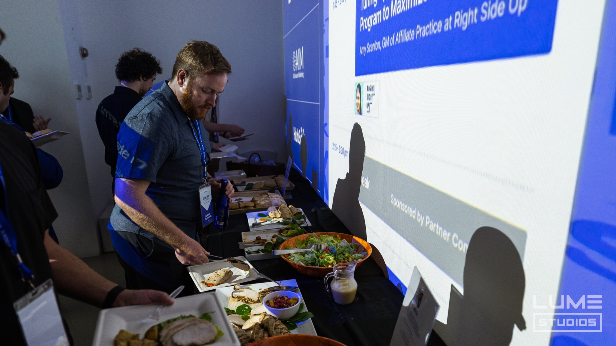 Bearded attendee with a lanyard fills a plate at a buffet in front of a blue session slide