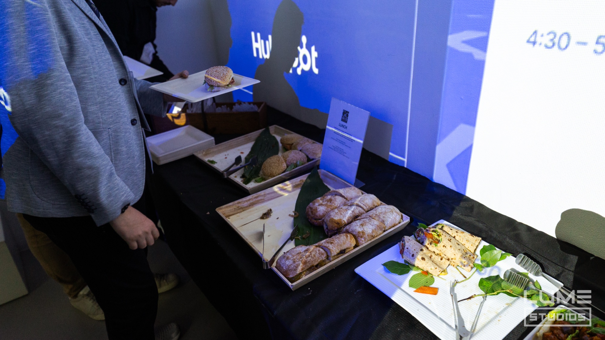 Trays of wrapped sandwiches and salad set out on a dark table before a blue projection wall