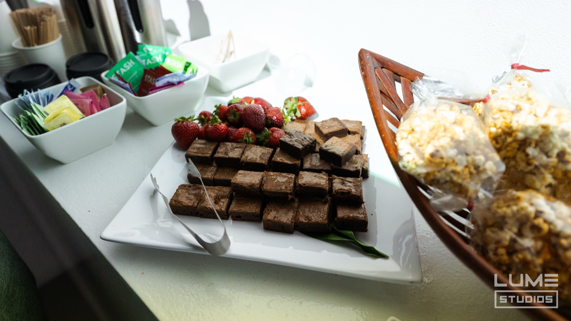 Platter of brownies and strawberries next to a basket of popcorn bags and tea bags on a white counter