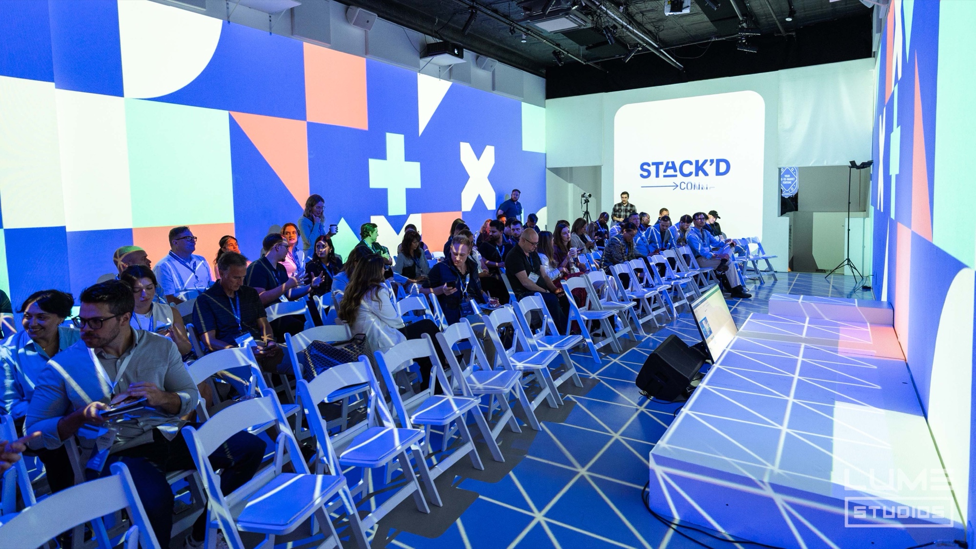 Audience in white folding chairs faces a Stack’d stage inside walls of blue and orange geometric shapes