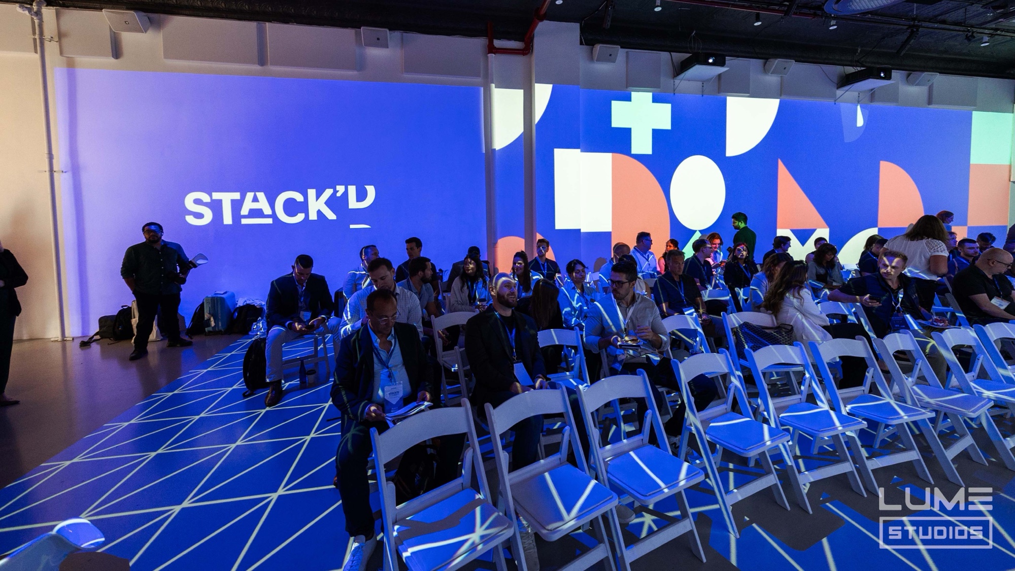Wide view of seated attendees in blue light as a Stack’d logo fills the projected wall at Lume Studios