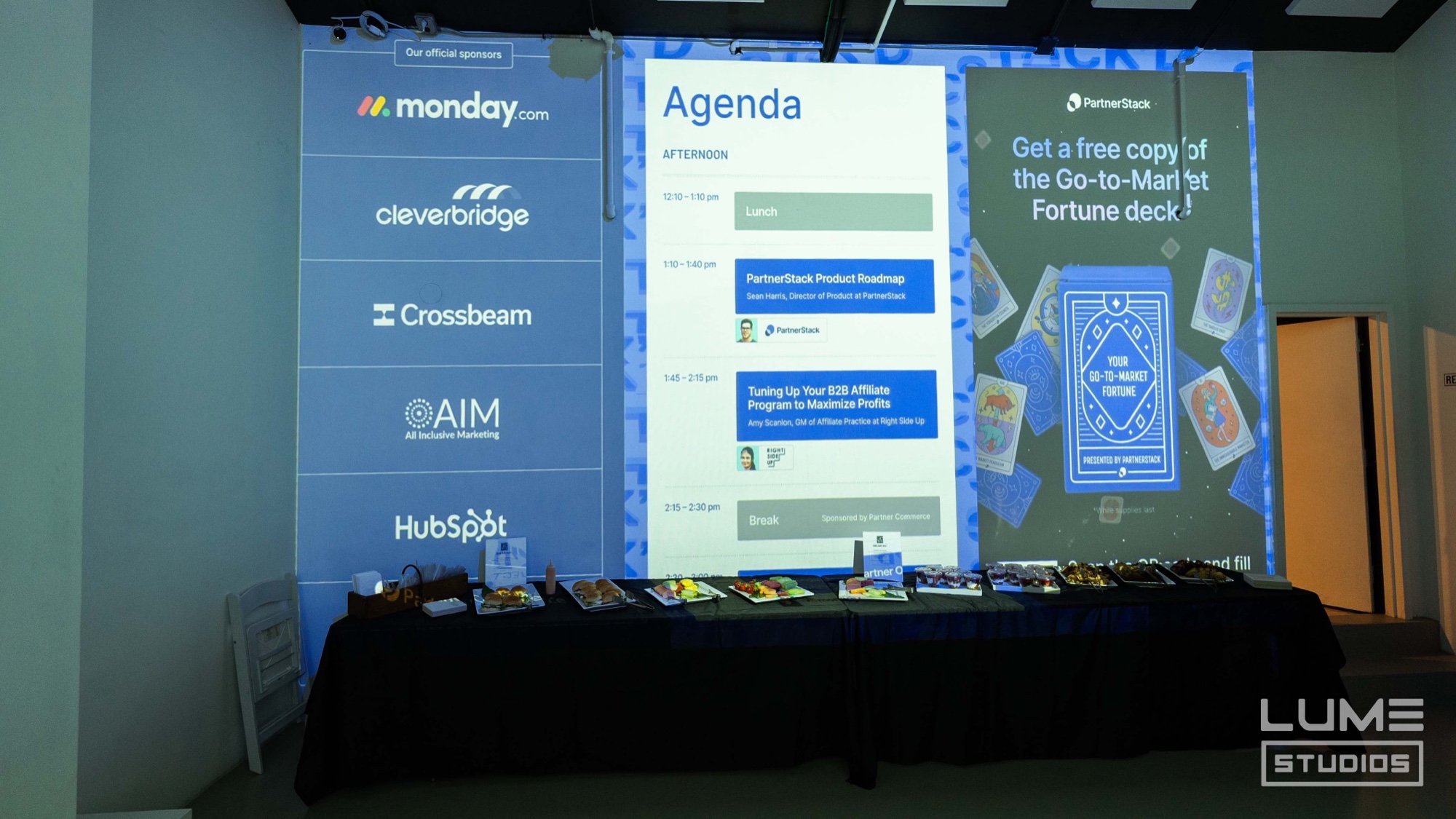 Projected agenda and sponsor logos above a table of printed materials in an empty blue lit room