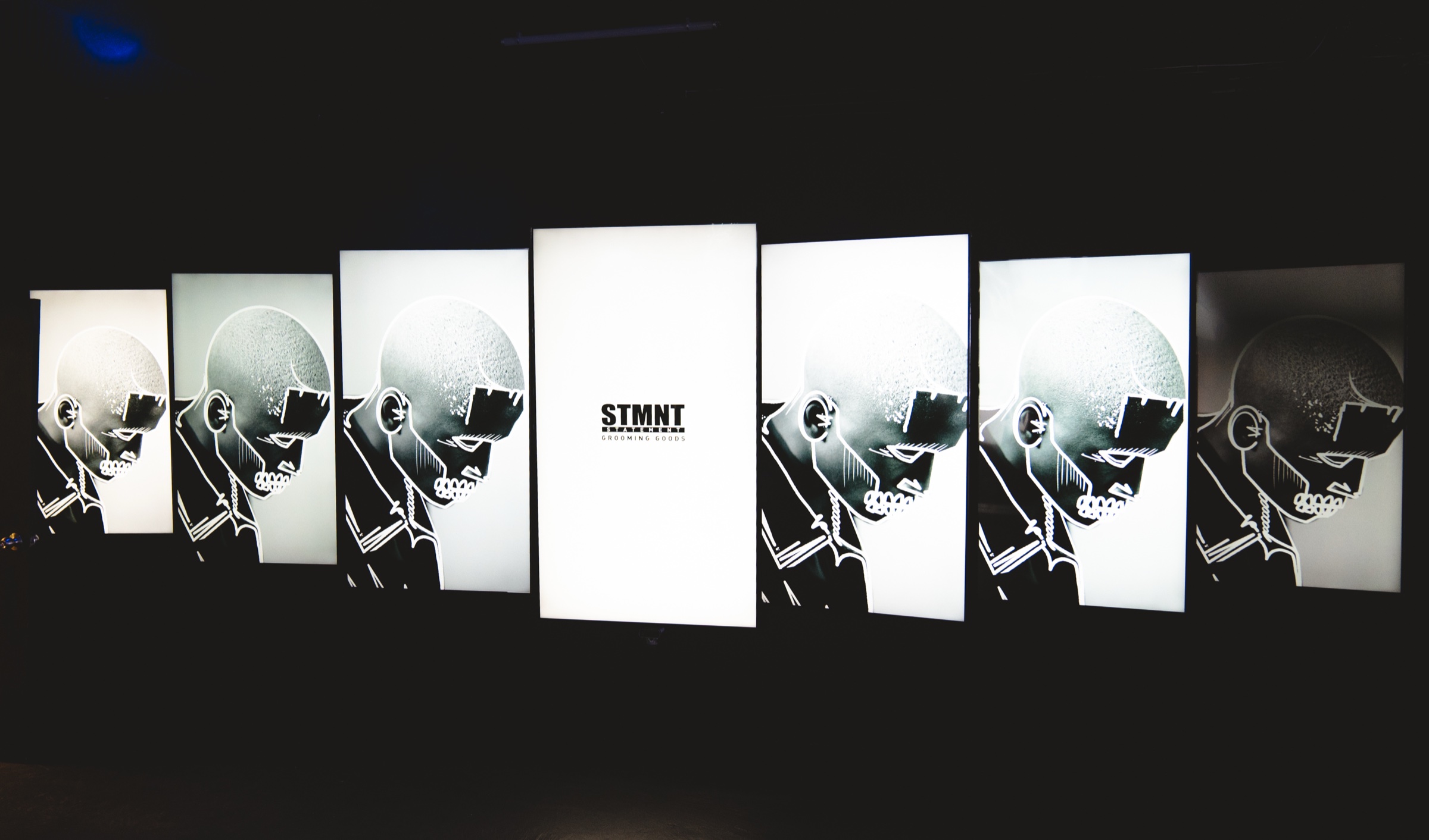 Seven lit panels in a dark room show black and white portraits of a shaved head beside the STMNT logo