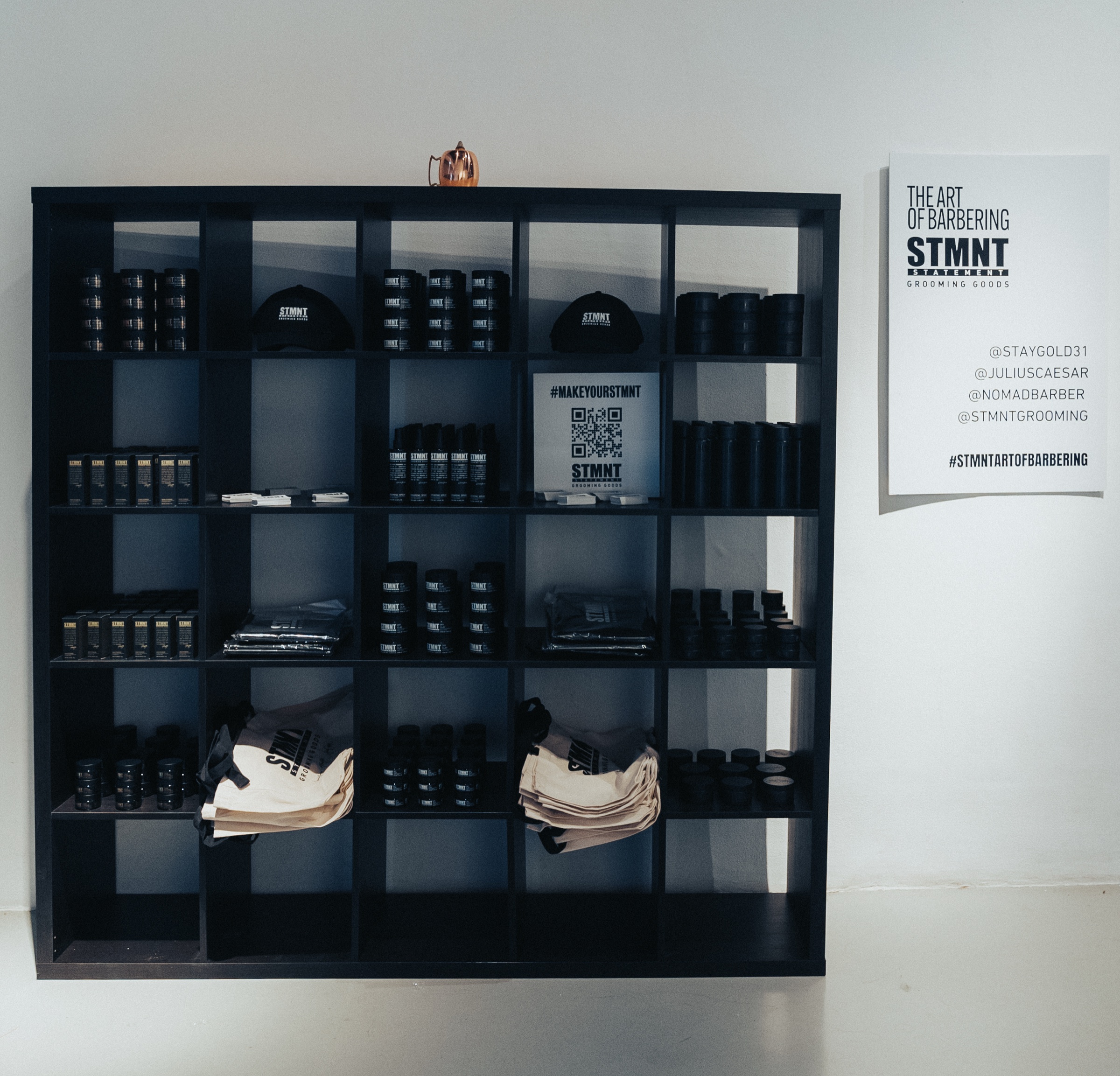 Dark shelving unit stocked with STMNT grooming products, caps and tote bags beside an event poster
