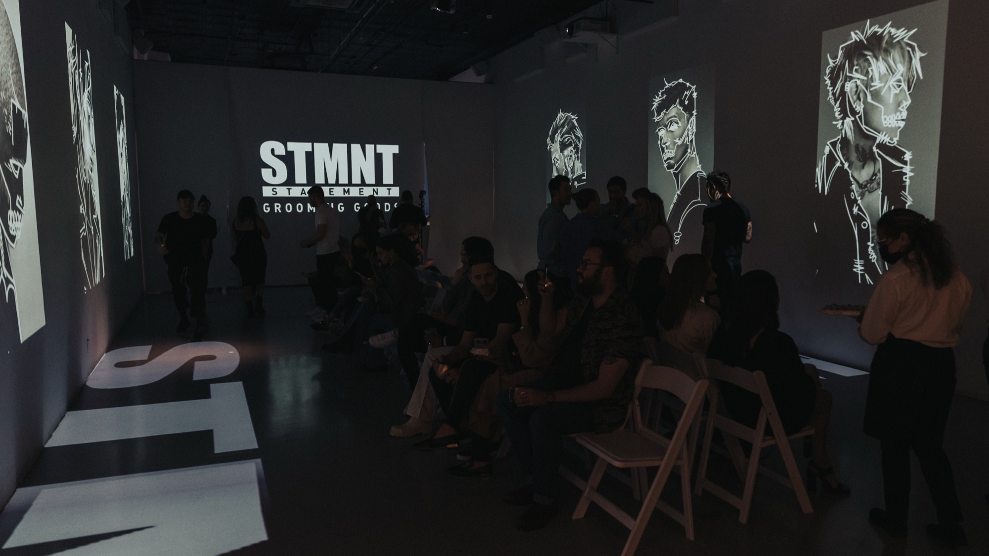 Seated guests in a dark room watching STMNT projections of sketched portraits in white on the walls