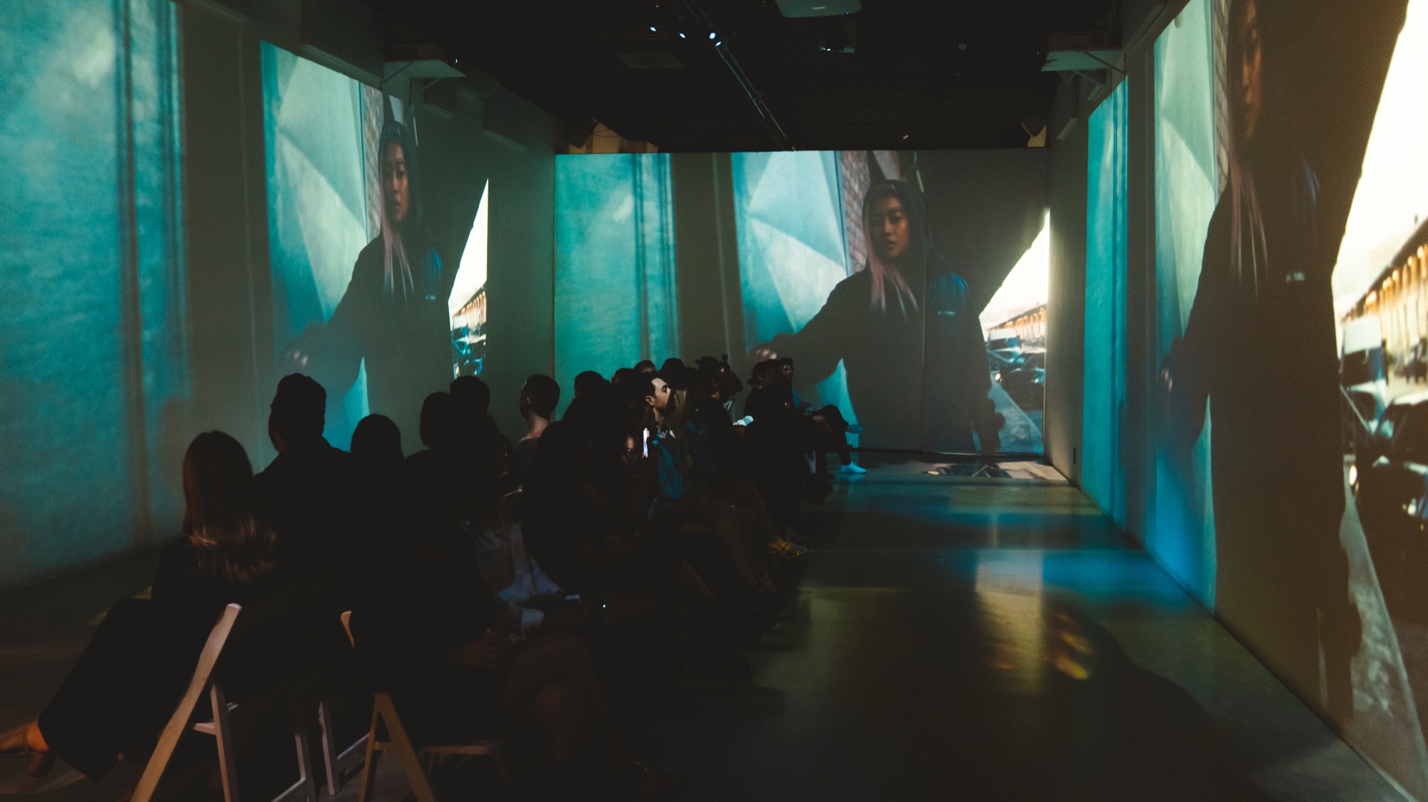 A seated audience watches teal tinted video of a person on the walls of a dark projection room