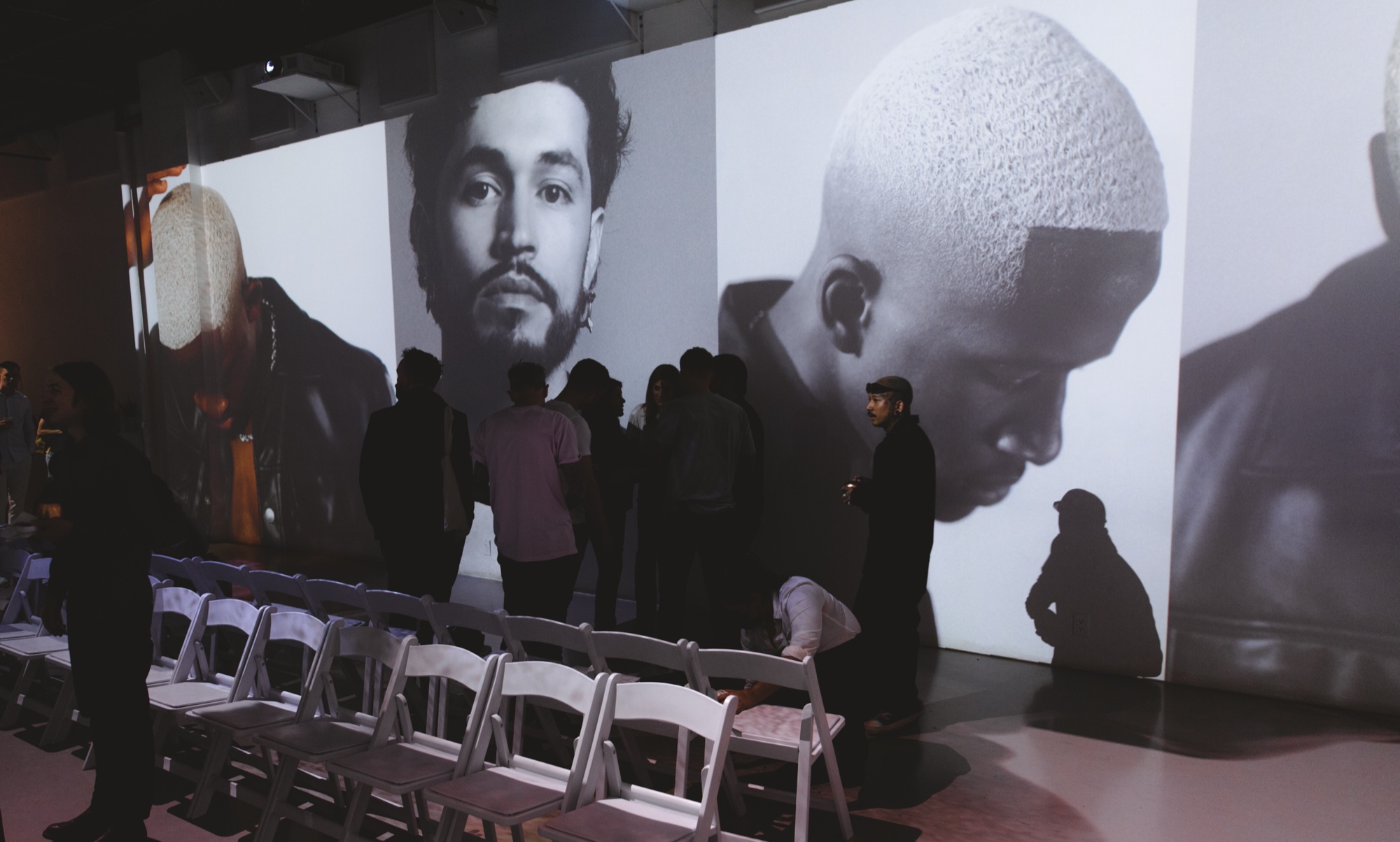 Rows of white folding chairs face walls projected with huge black and white portraits of men with short hair