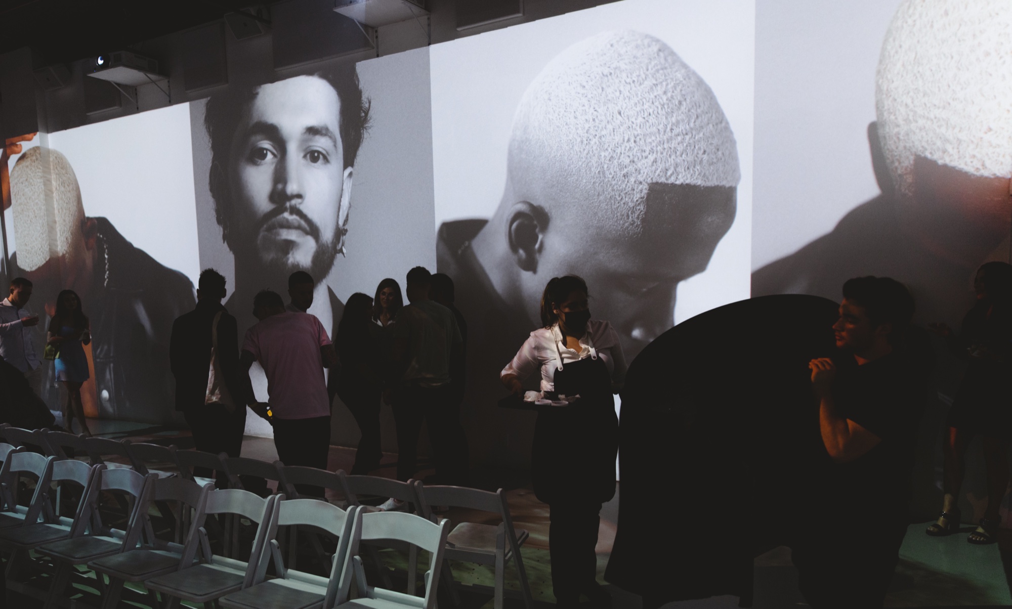 Guests stand before large black and white projected portraits of men with a row of white chairs in front