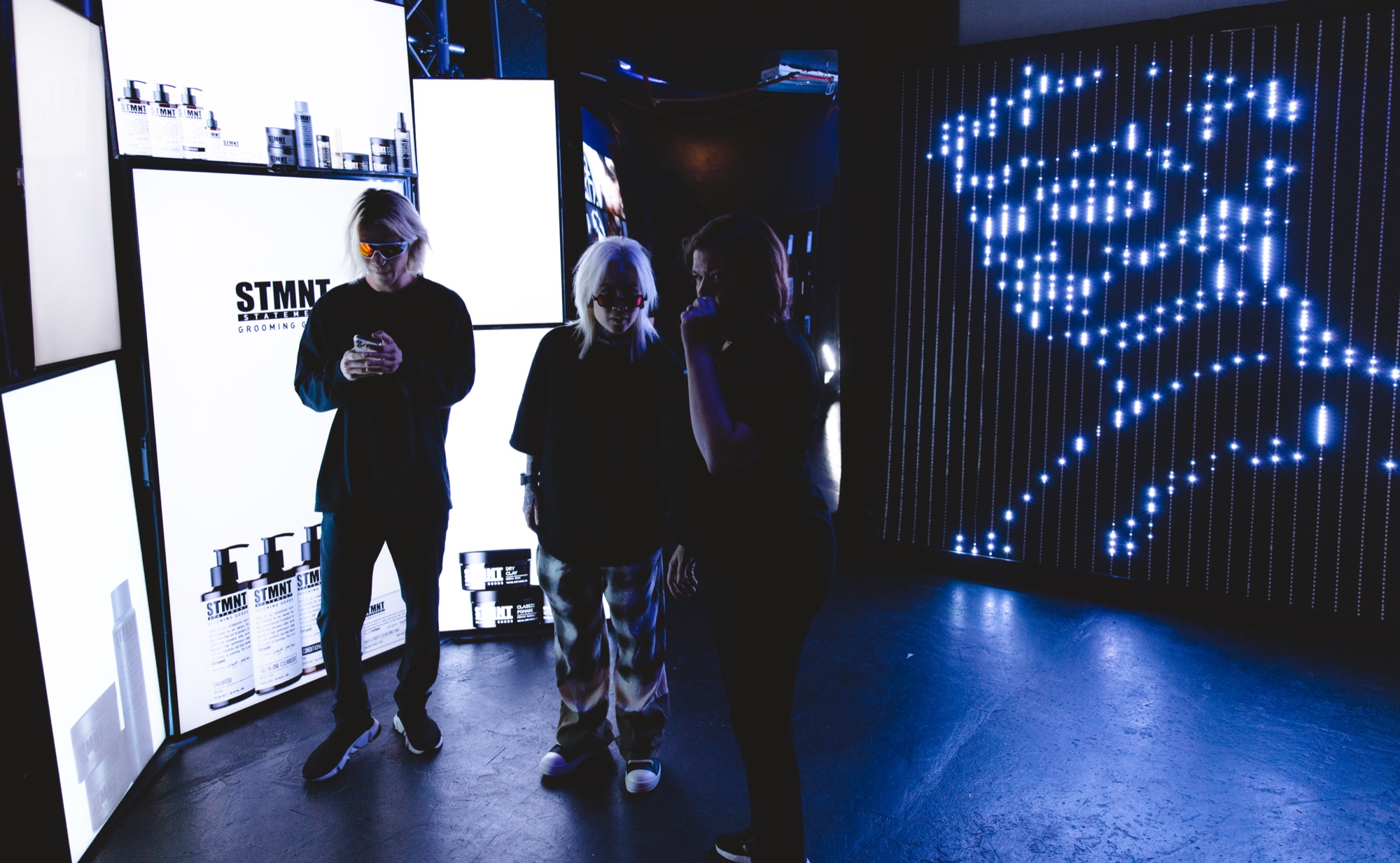 Three guests stand between backlit STMNT product panels and a wall of blue LED dots forming a face