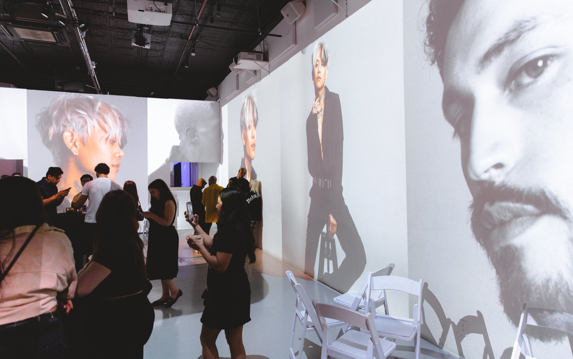 Guests photograph huge black and white portraits projected on the walls of a white room with folding chairs