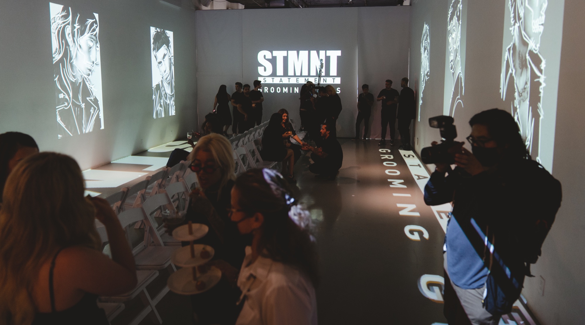 Guests seated at white tables under a projected STMNT logo as a photographer shoots from the aisle