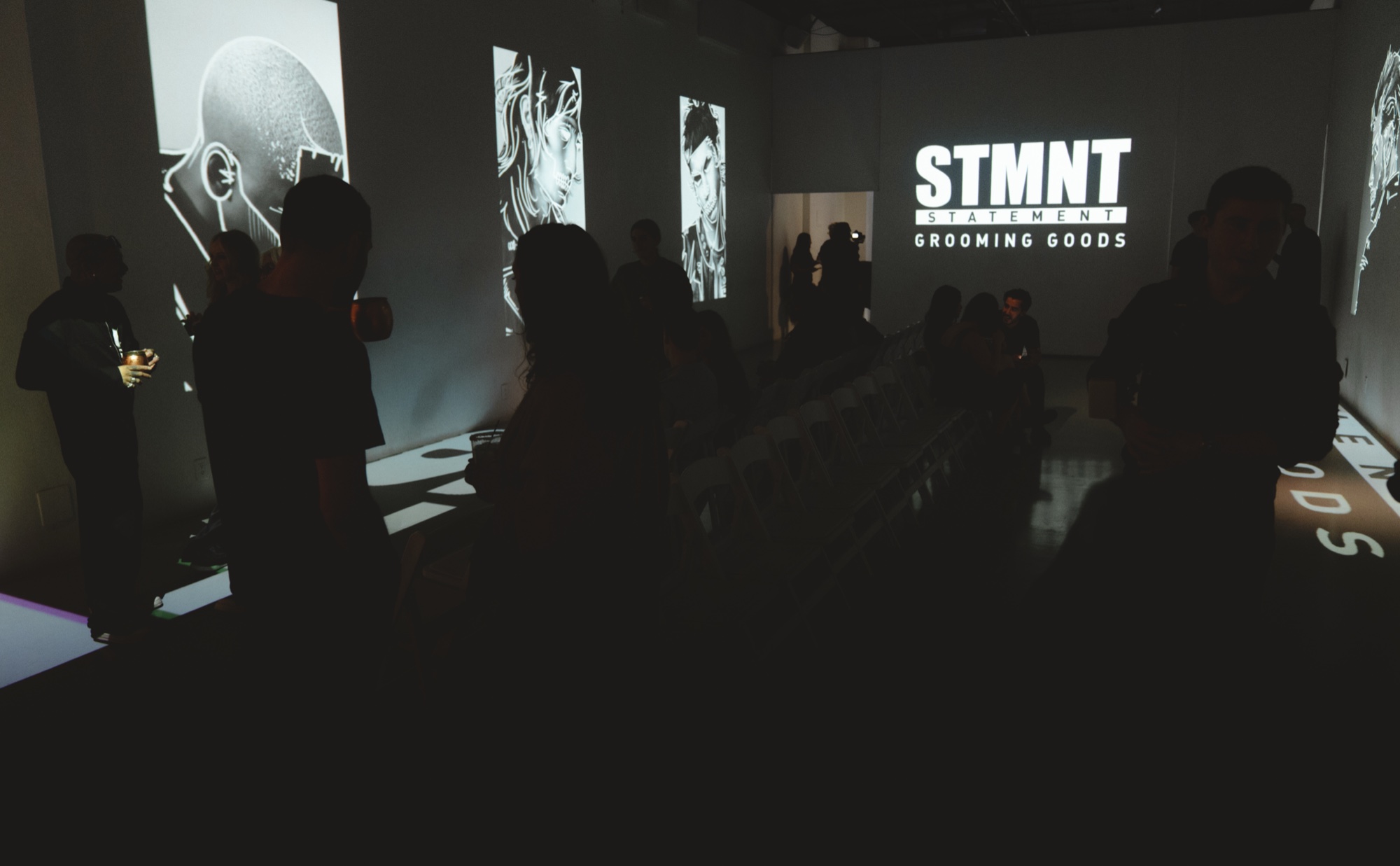 Silhouetted guests in a dark room lit by a STMNT Grooming Goods projection and portrait sketches