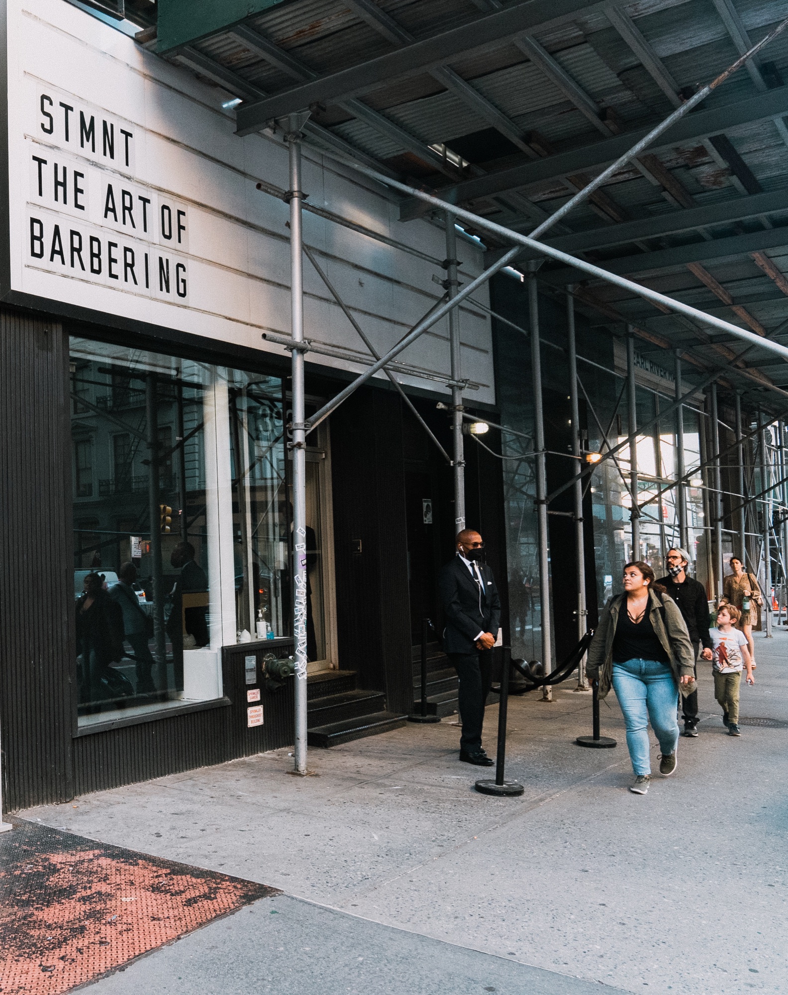 A marquee reads STMNT The Art of Barbering under scaffolding as a doorman stands by a velvet rope