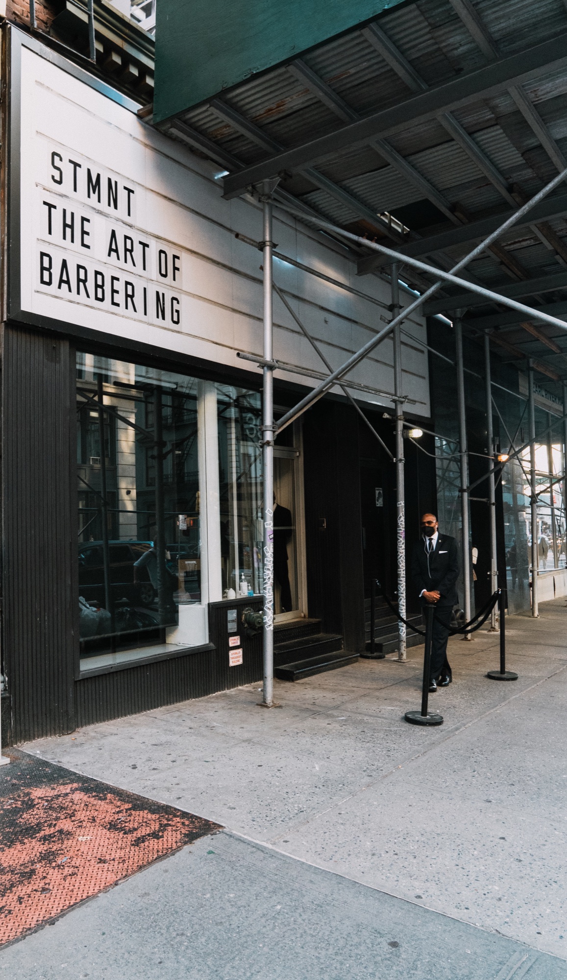 Storefront marquee reading STMNT The Art of Barbering under scaffolding, a suited doorman by a rope