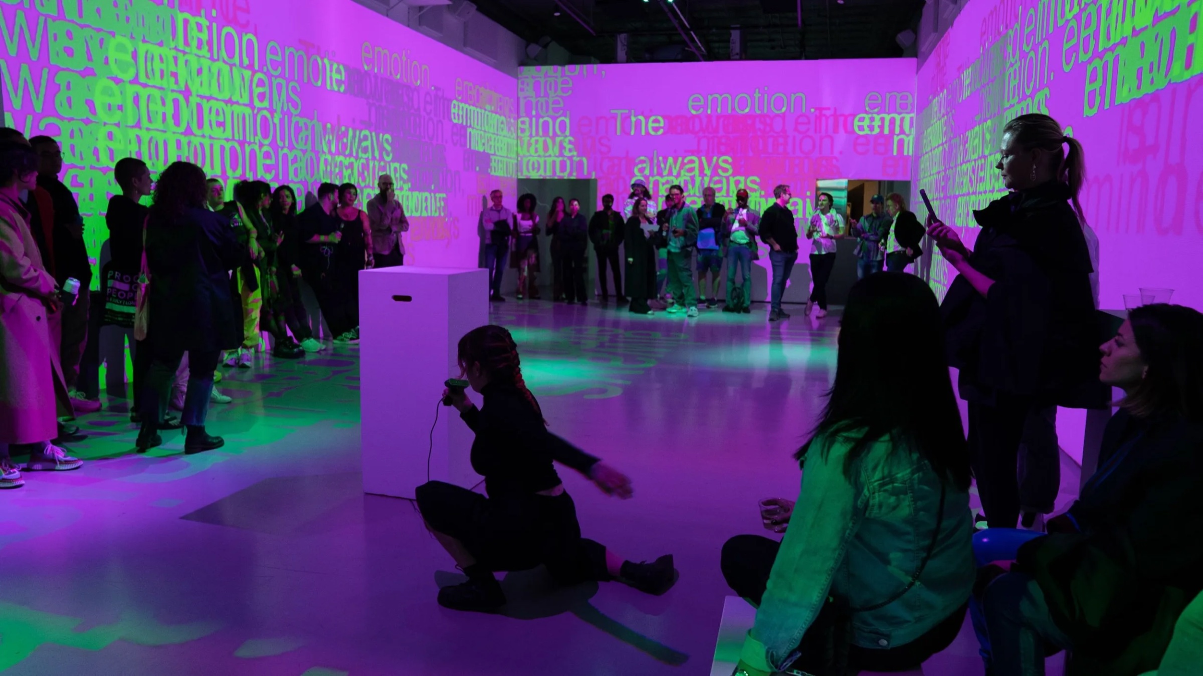A performer kneels with a microphone on the floor of the magenta immersive room while the crowd watches from the walls