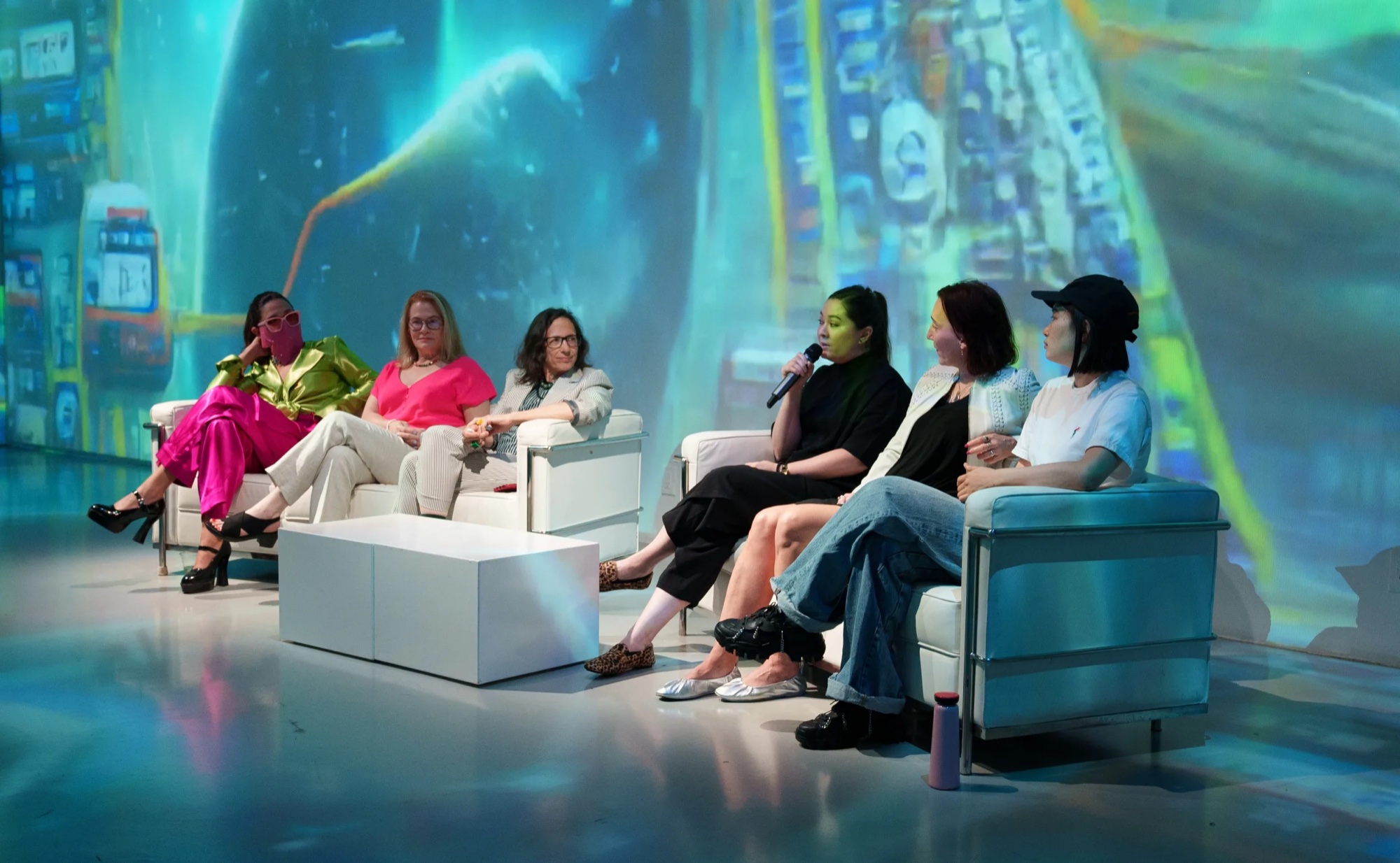 Six speakers sit in white lounge chairs on a stage washed in blue and green projection during a Subjective Art Festival panel
