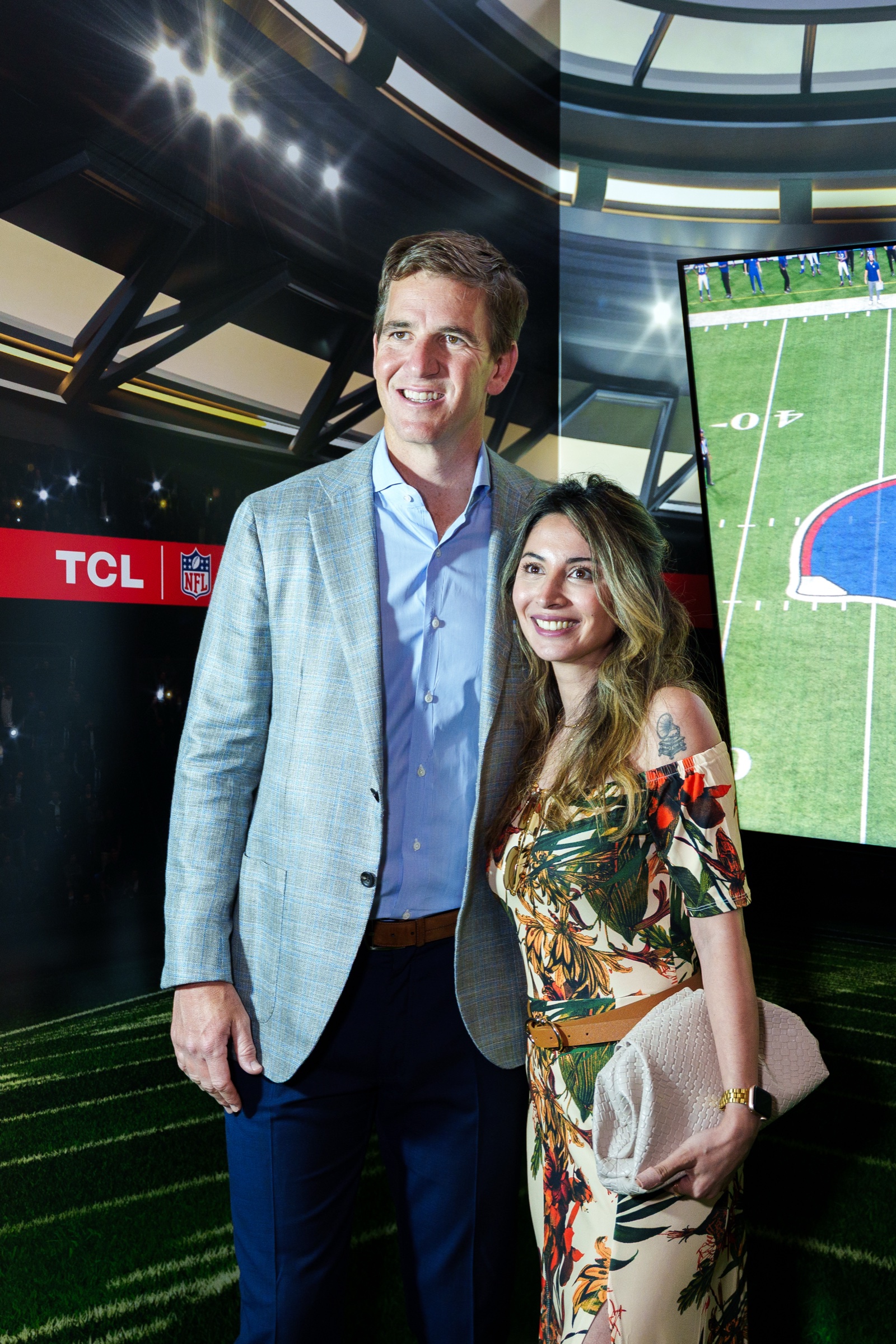 Man in a grey blazer and woman in a floral dress pose before a projected stadium and TCL NFL logo
