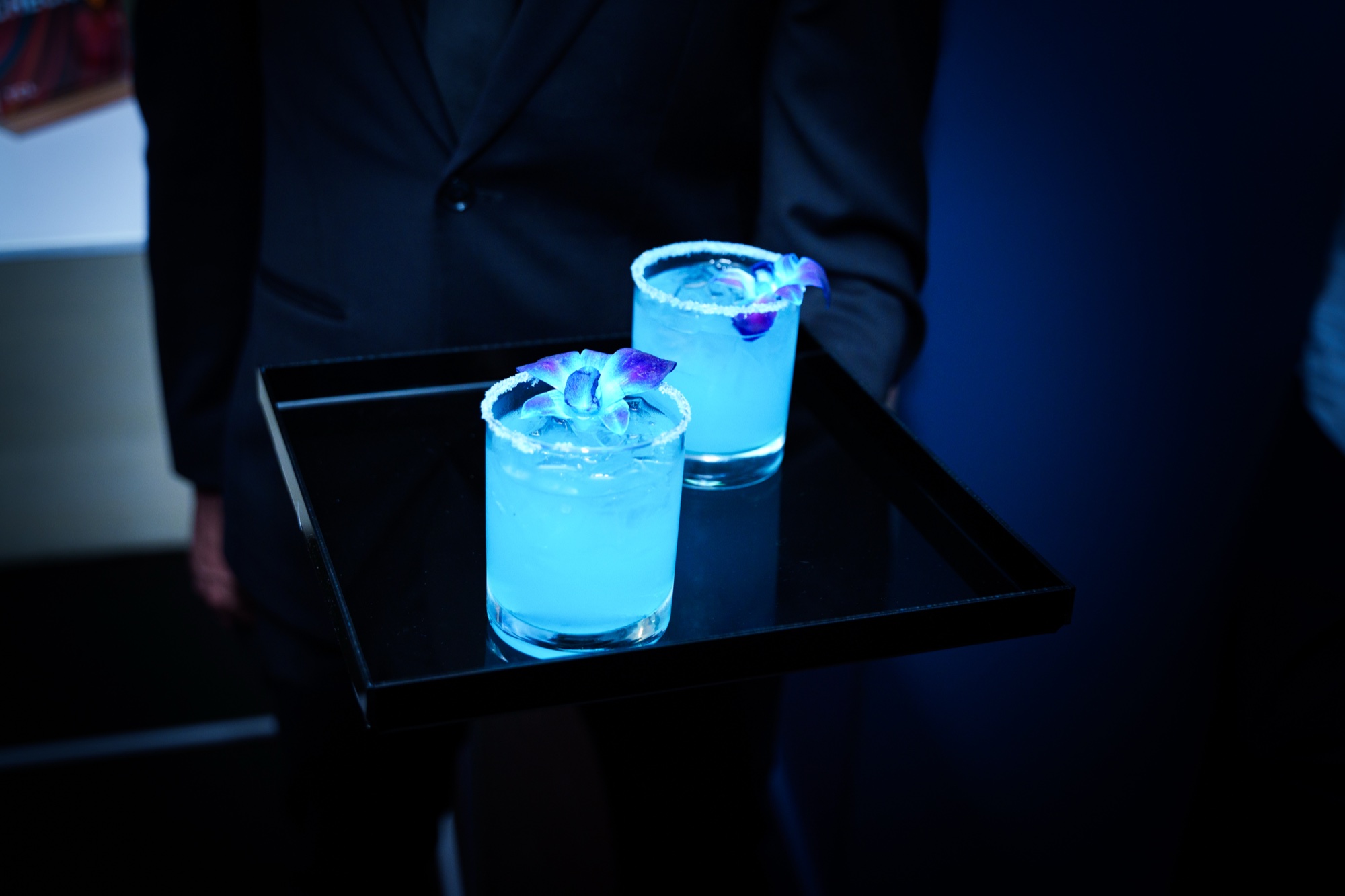 Server holds a black tray with two glowing blue cocktails garnished with purple orchids