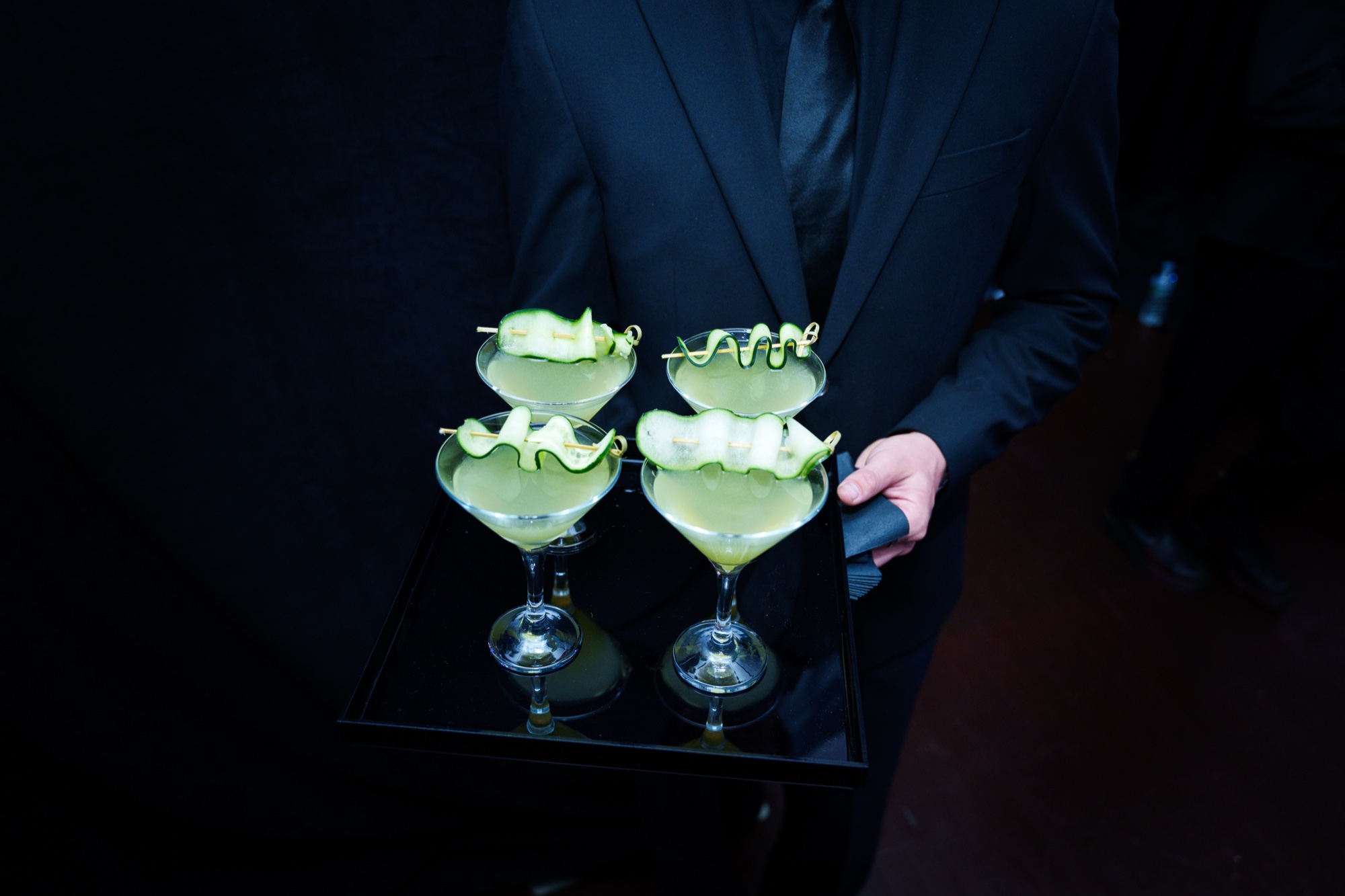Server in a dark suit holds a black tray of four cocktails garnished with cucumber ribbons