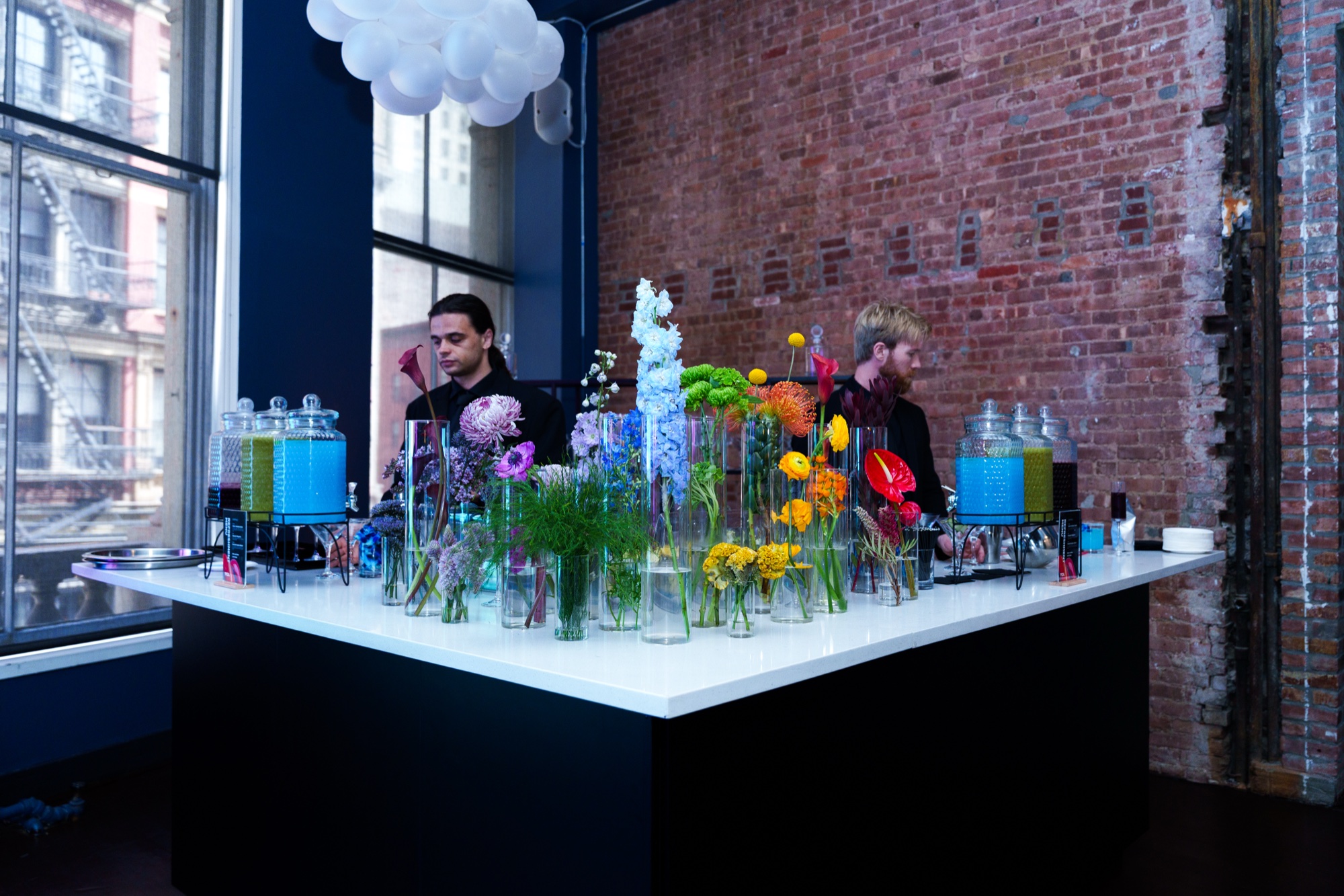 Two bartenders behind a black bar crowded with colourful flowers and drink dispensers under a cloud lamp
