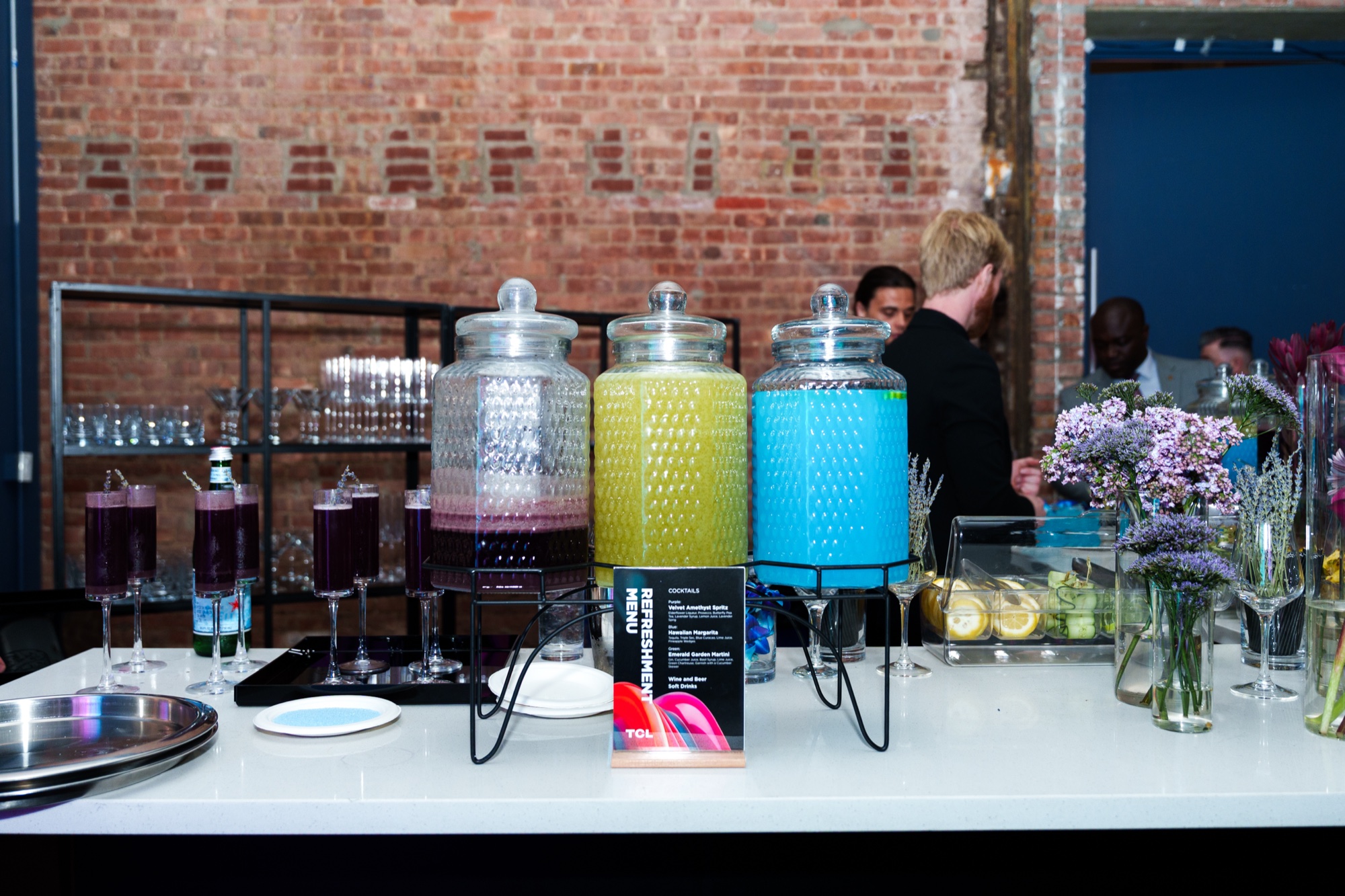 Three glass dispensers of purple, yellow and blue drinks on a white bar before an exposed brick wall