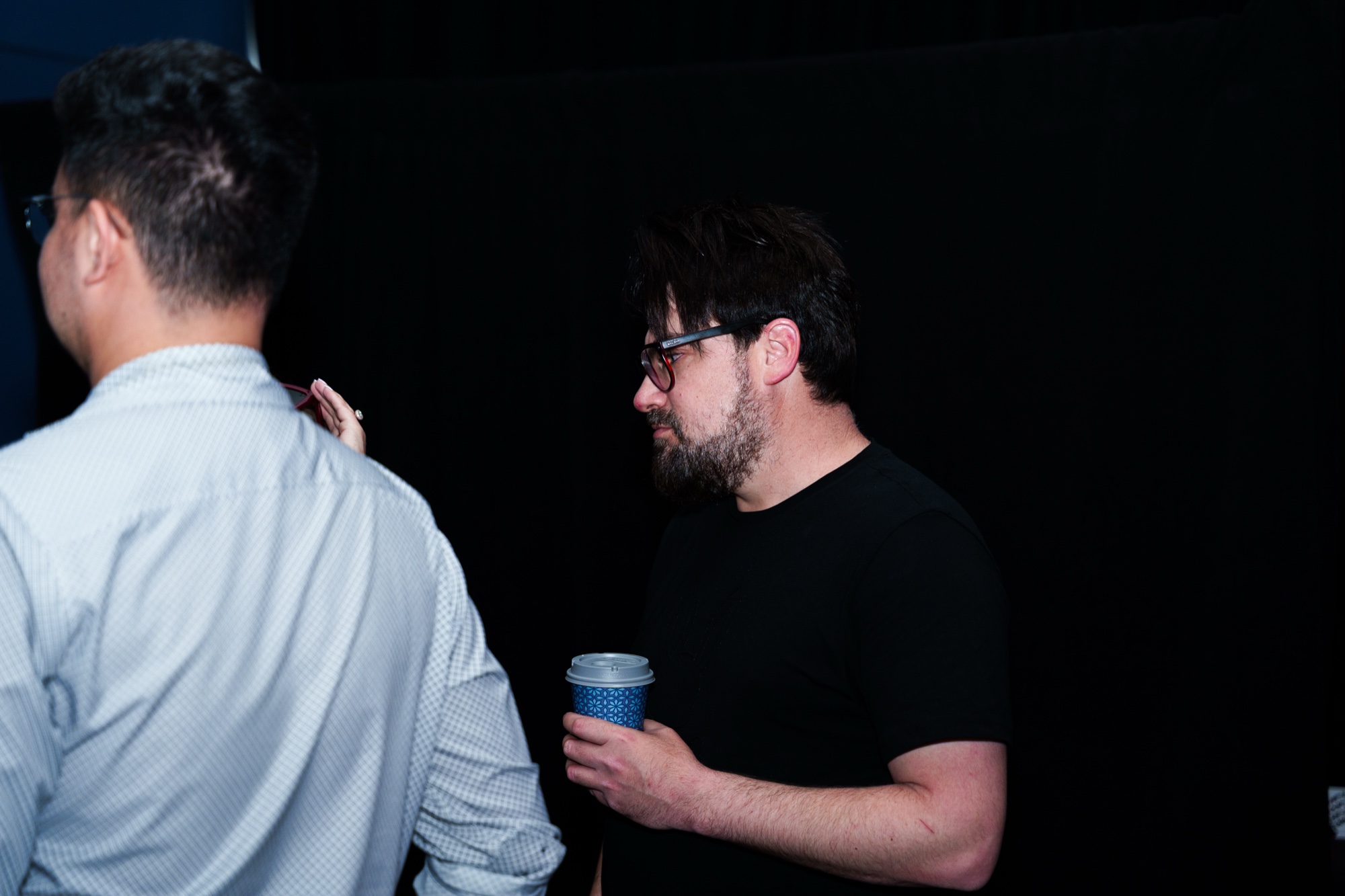 Bearded man in glasses and a black tee holds a blue cup beside a man in a white shirt