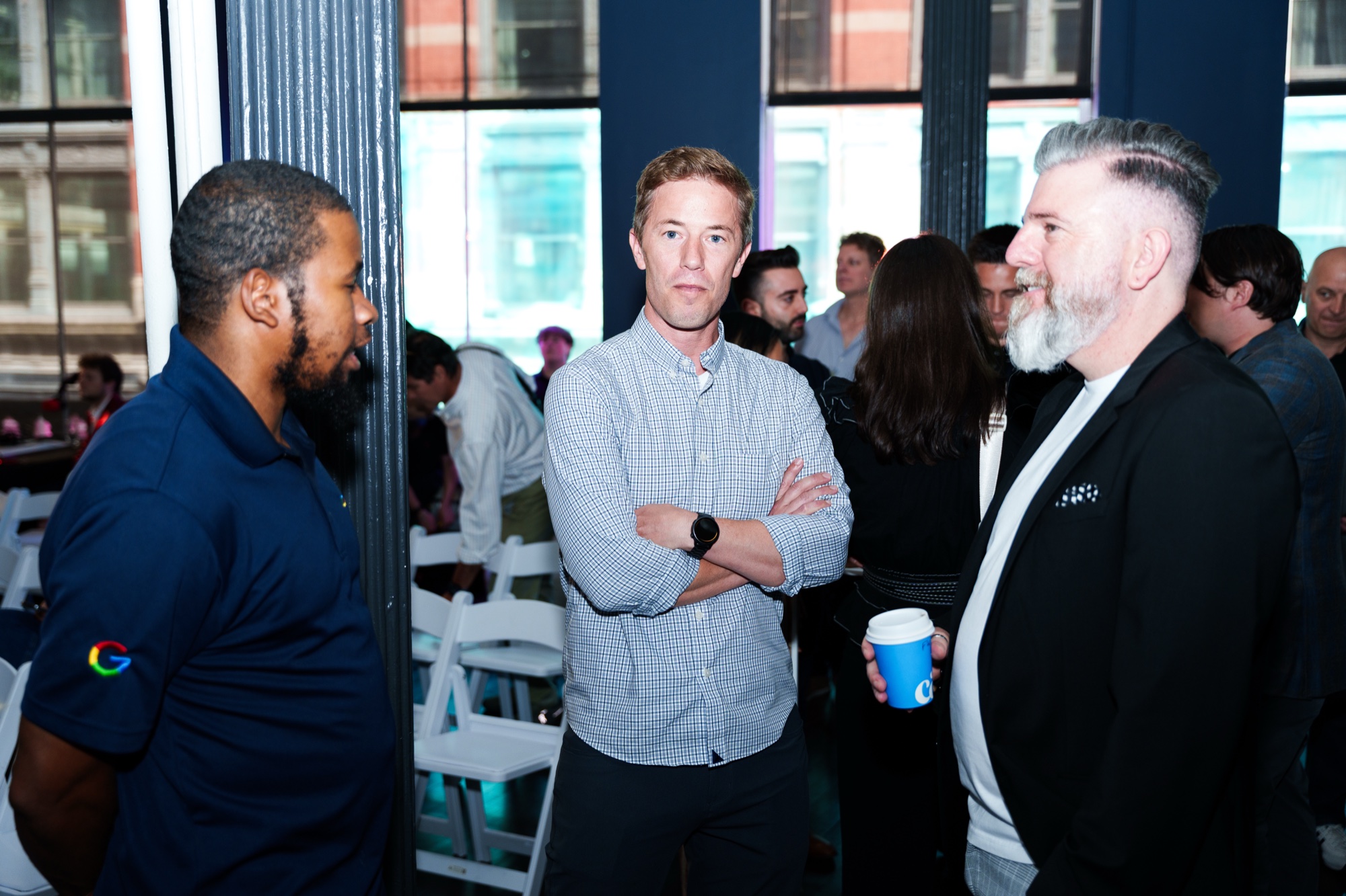 Three men chat by a window at a daytime event, one holding a coffee cup