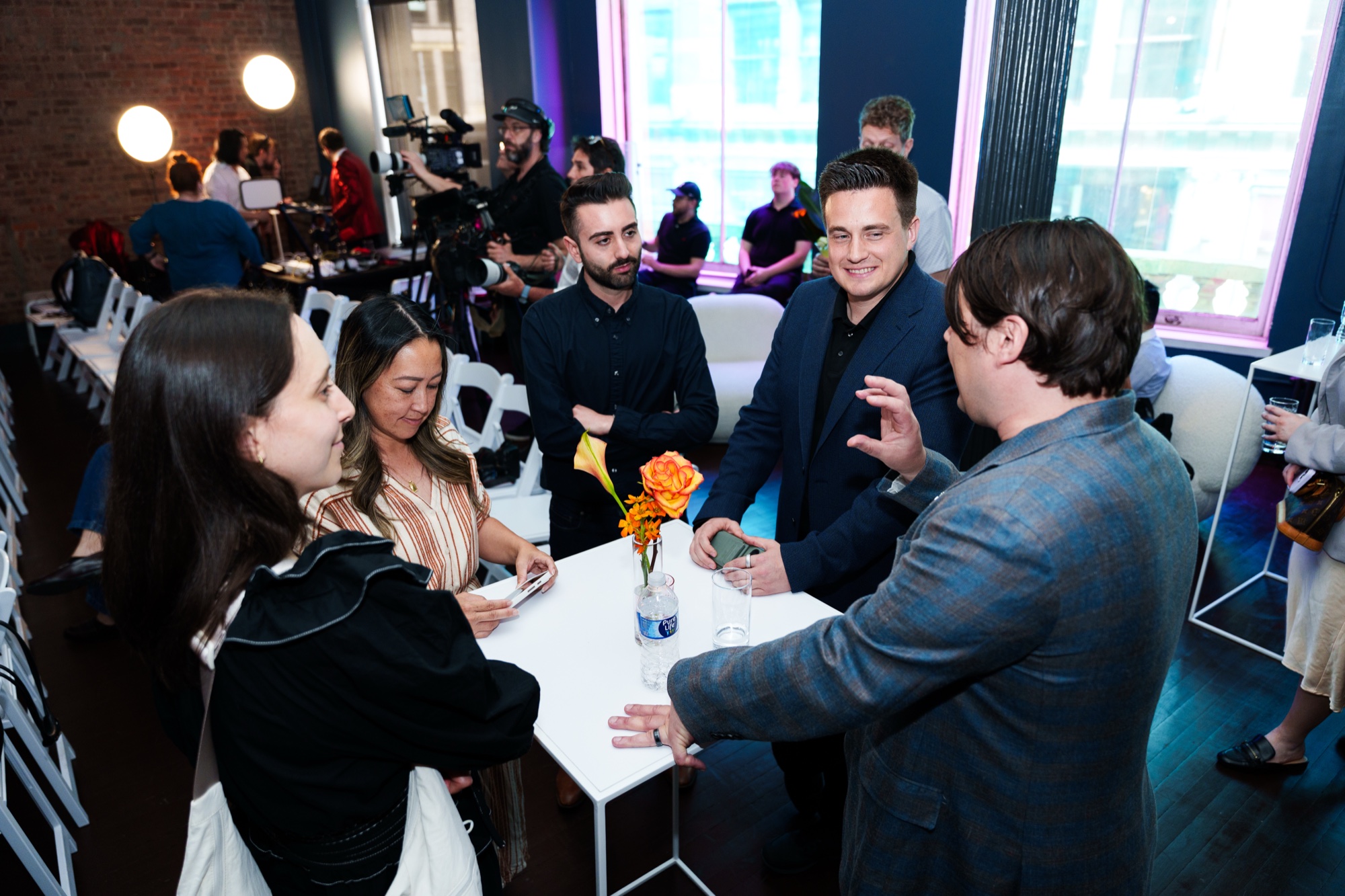 Guests chat around a white cocktail table with an orange flower while a camera crew films behind them