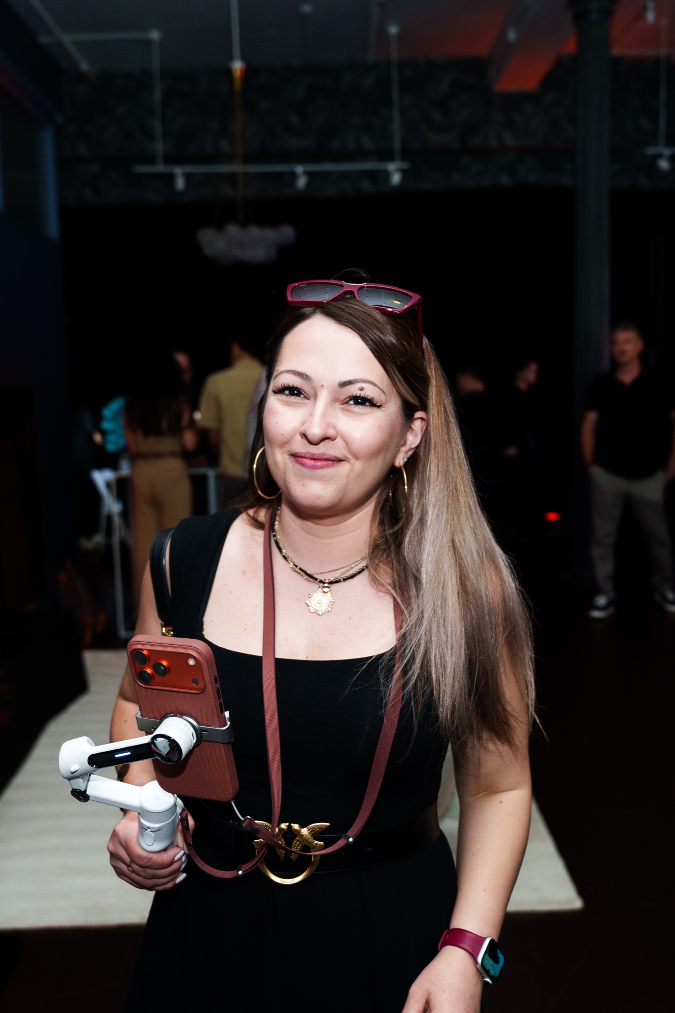 Smiling woman in a black dress with sunglasses on her head holds a phone on a gimbal in a loft