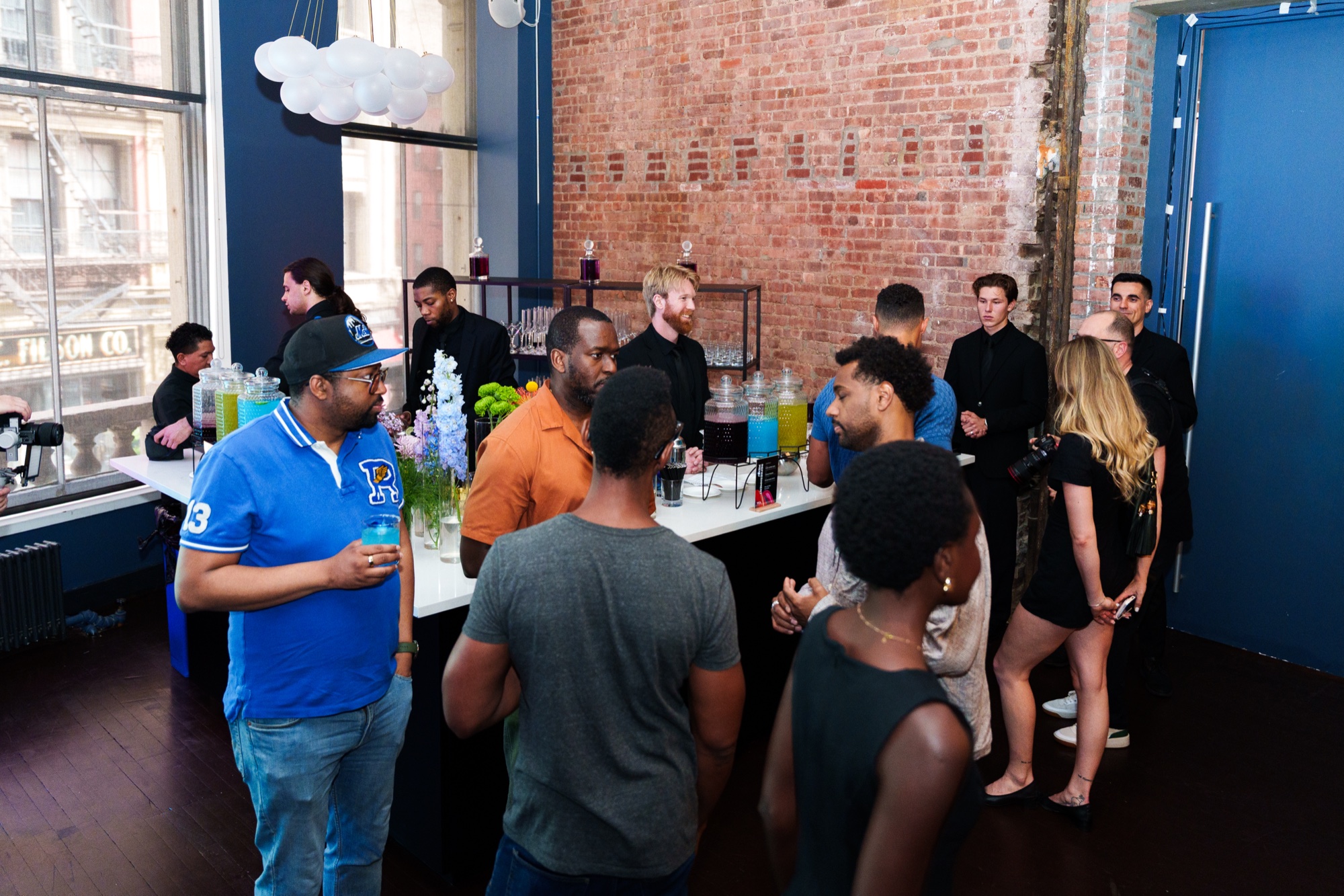 Guests mingle by a bar with flowers and drink dispensers in a brick loft with big windows