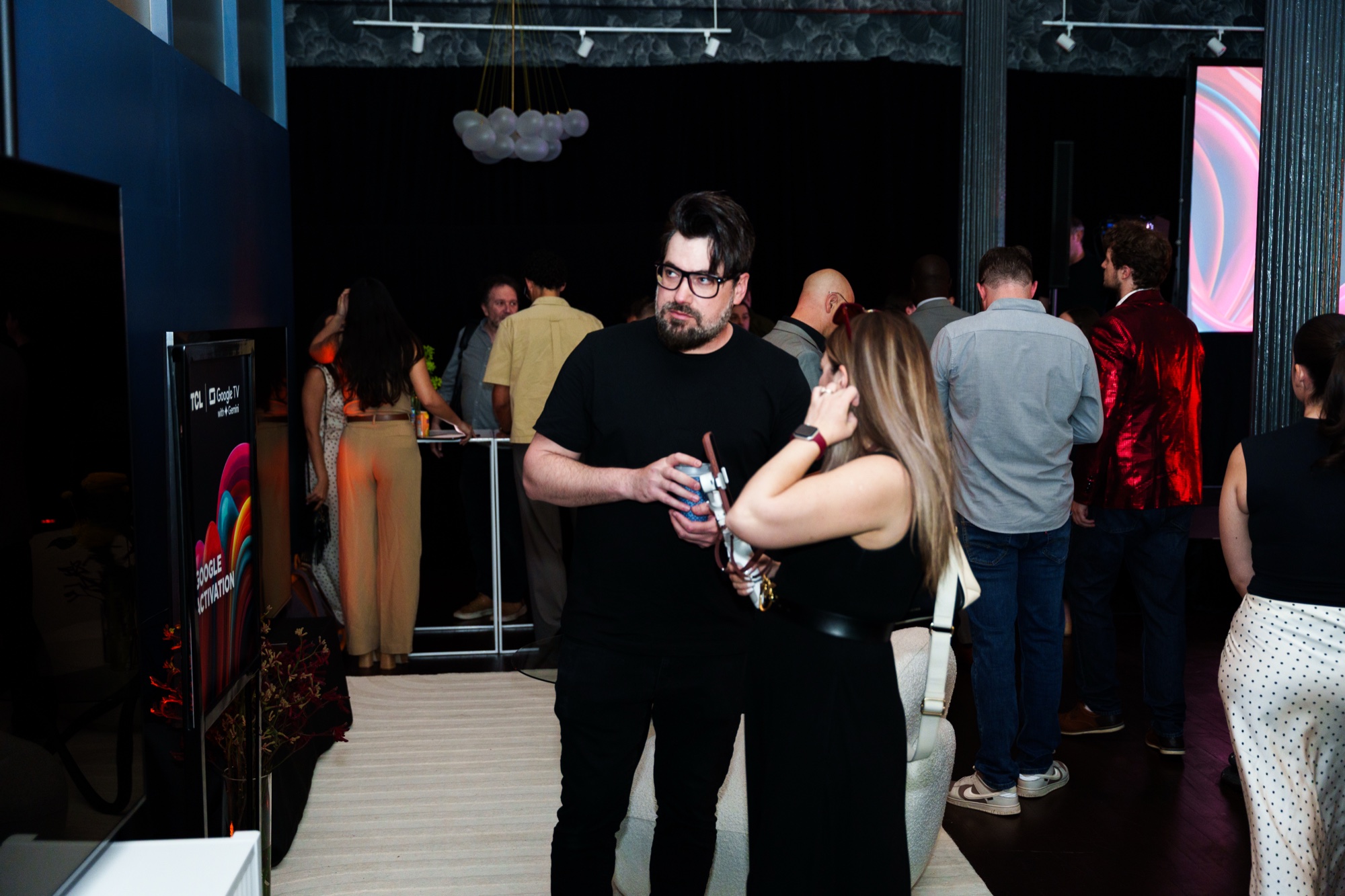 Couple in black talk in a dim lounge with a bubble chandelier and guests in the background