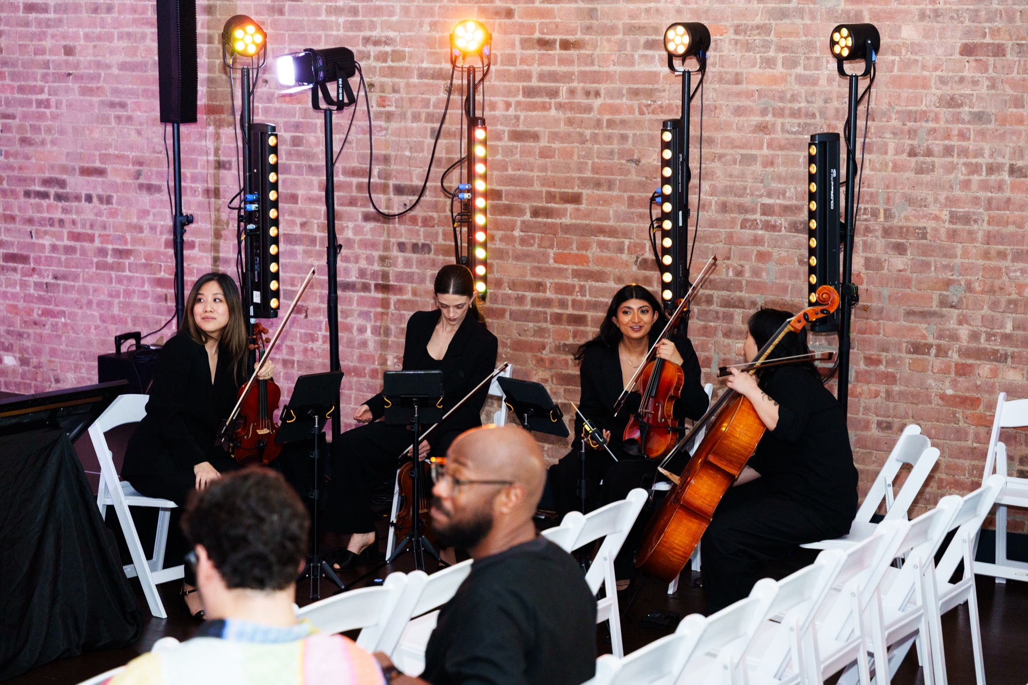 String quartet in black performs before a brick wall between lit stands as guests sit on white chairs