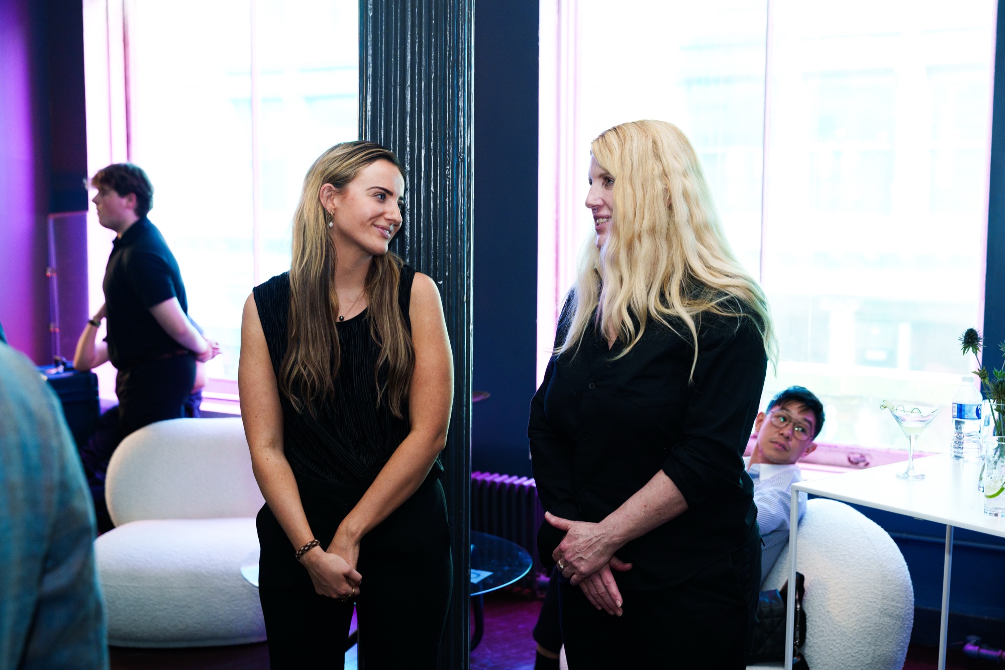 Two women in black talk beside a white curved sofa in a bright loft with purple accents